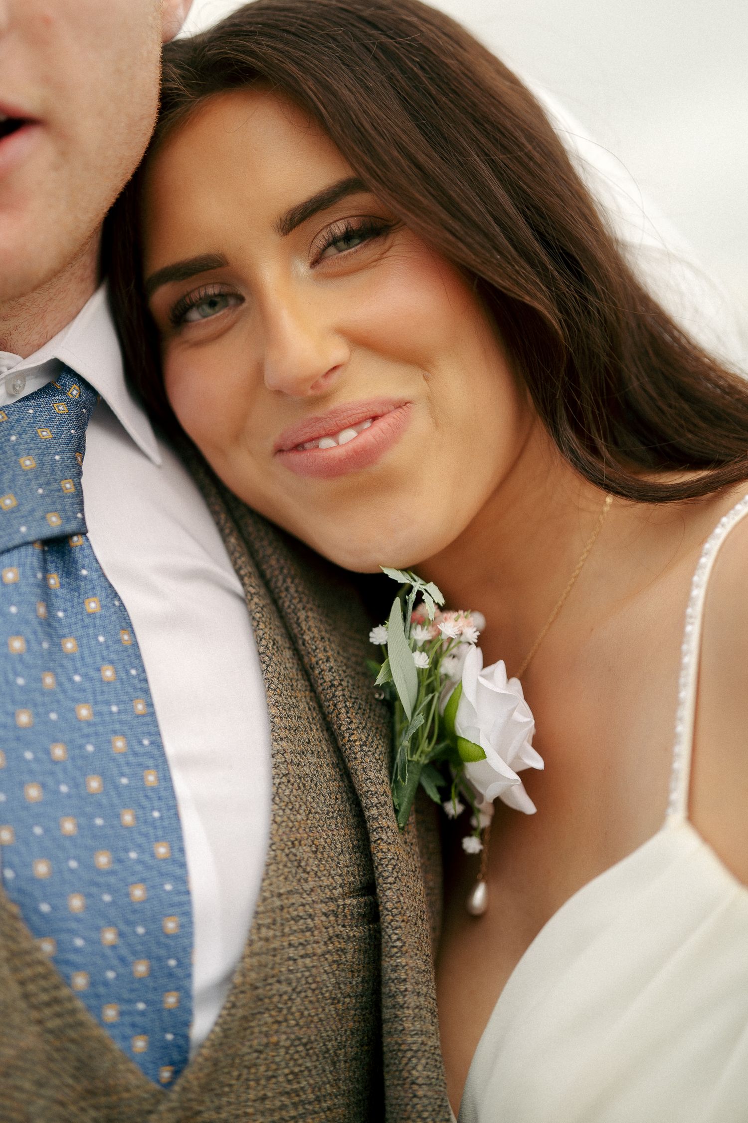 Close-up of a smiling bride in a white dress resting her head on the groom's shoulder, a floral boutonniere pinned to his tweed jacket.