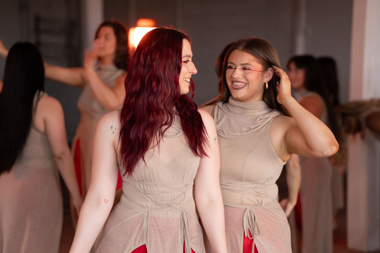 Two smiling women in matching beige halter dresses with red accents laugh together during a group dance rehearsal in a bright studio.