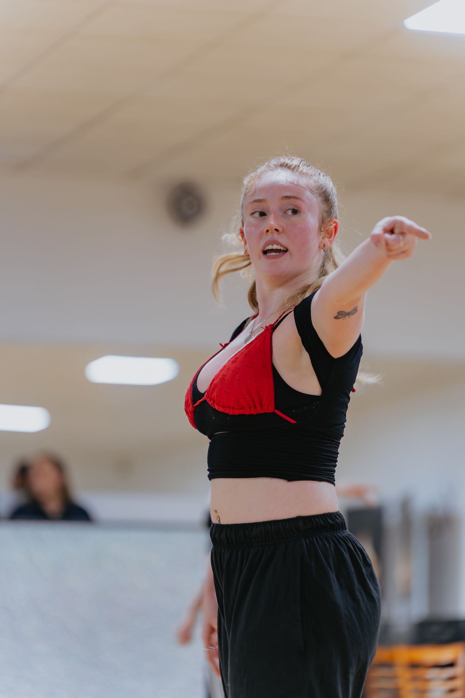 Young woman with blonde hair pulled back points outward while teaching a dance class in a bright studio, wearing a black and red top.