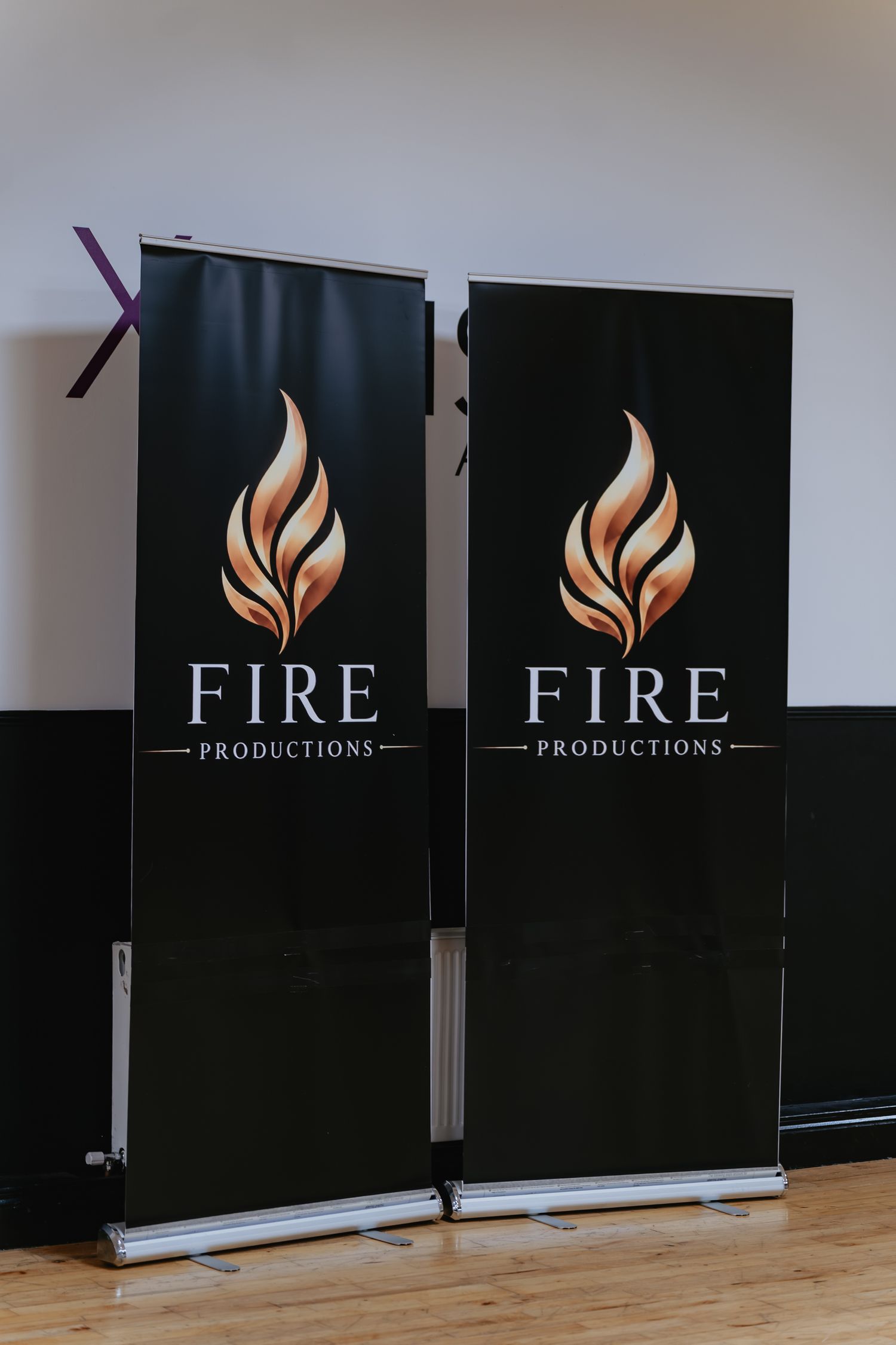 Two black Fire Productions roll-up banners with an orange flame logo stand side by side on a wooden floor against a white wall.