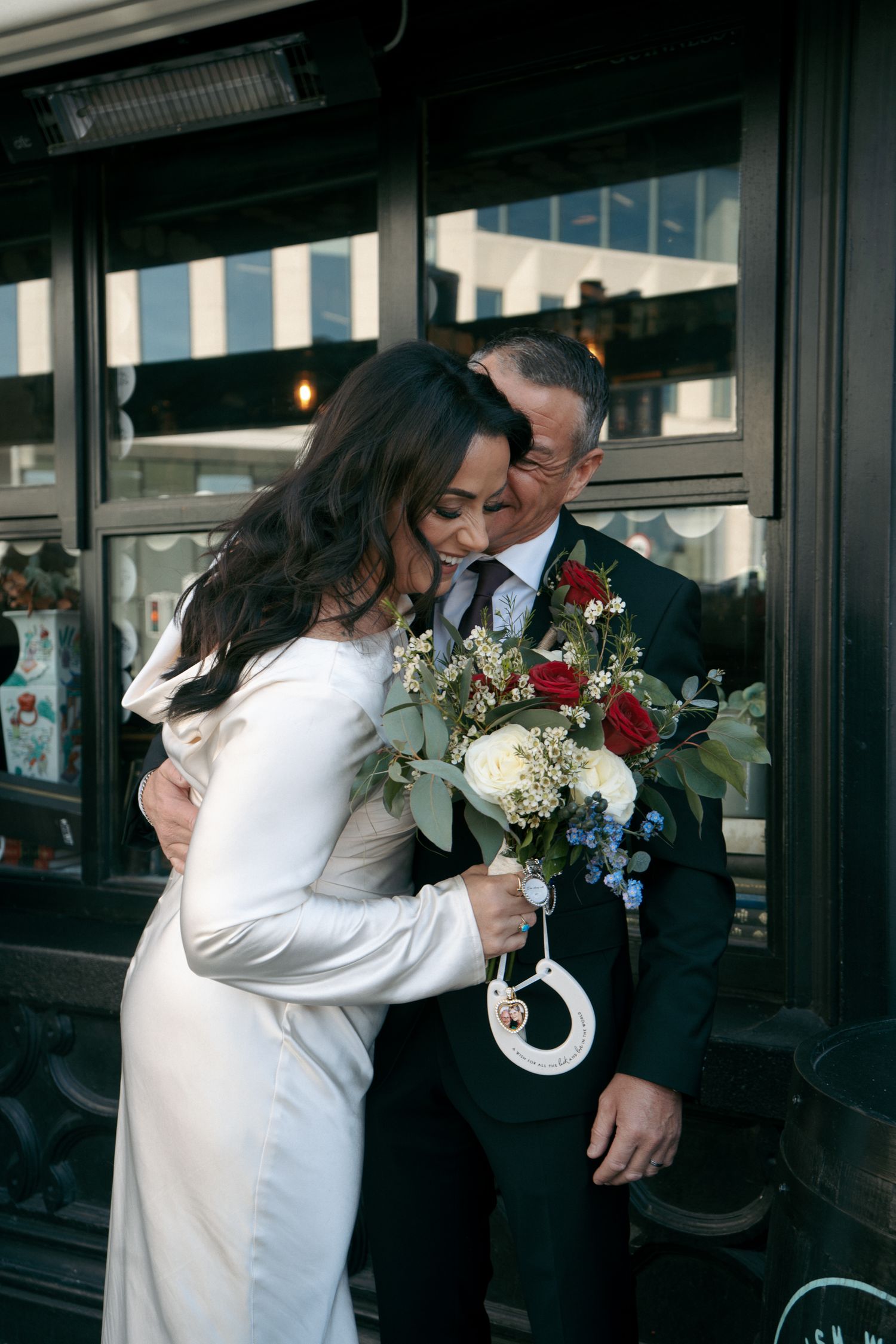 Bride in a white satin dress embraces her smiling groom outside a dark storefront, holding a bouquet of red, white and blue flowers.