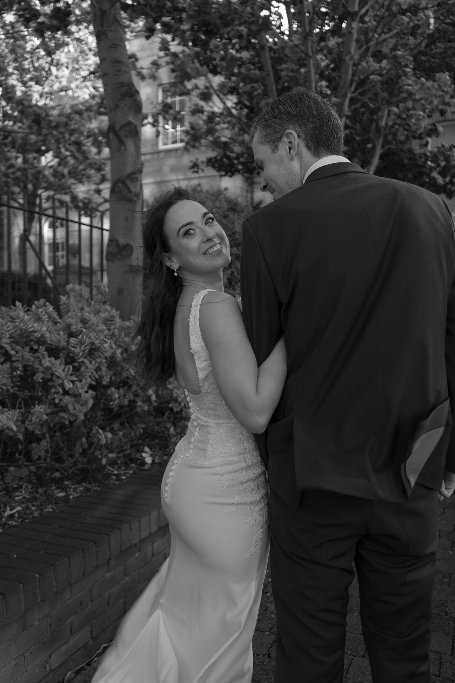 Black and white wedding portrait of a smiling bride in a fitted satin gown glancing over her shoulder while holding her groom's arm outdoors