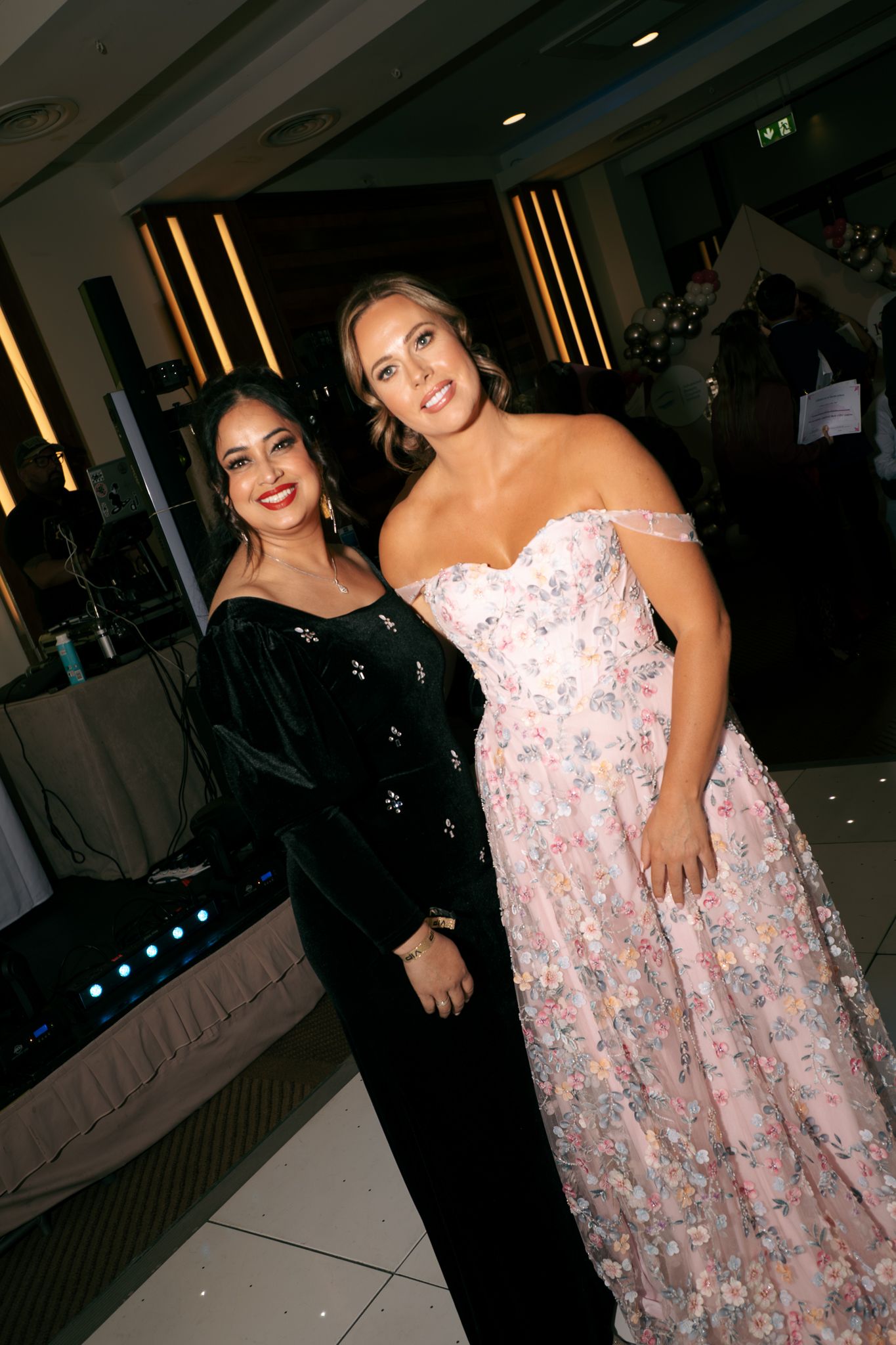 Two women in formal evening gowns, one in black velvet and one in a pink floral off-shoulder dress, pose together at an elegant event venue.