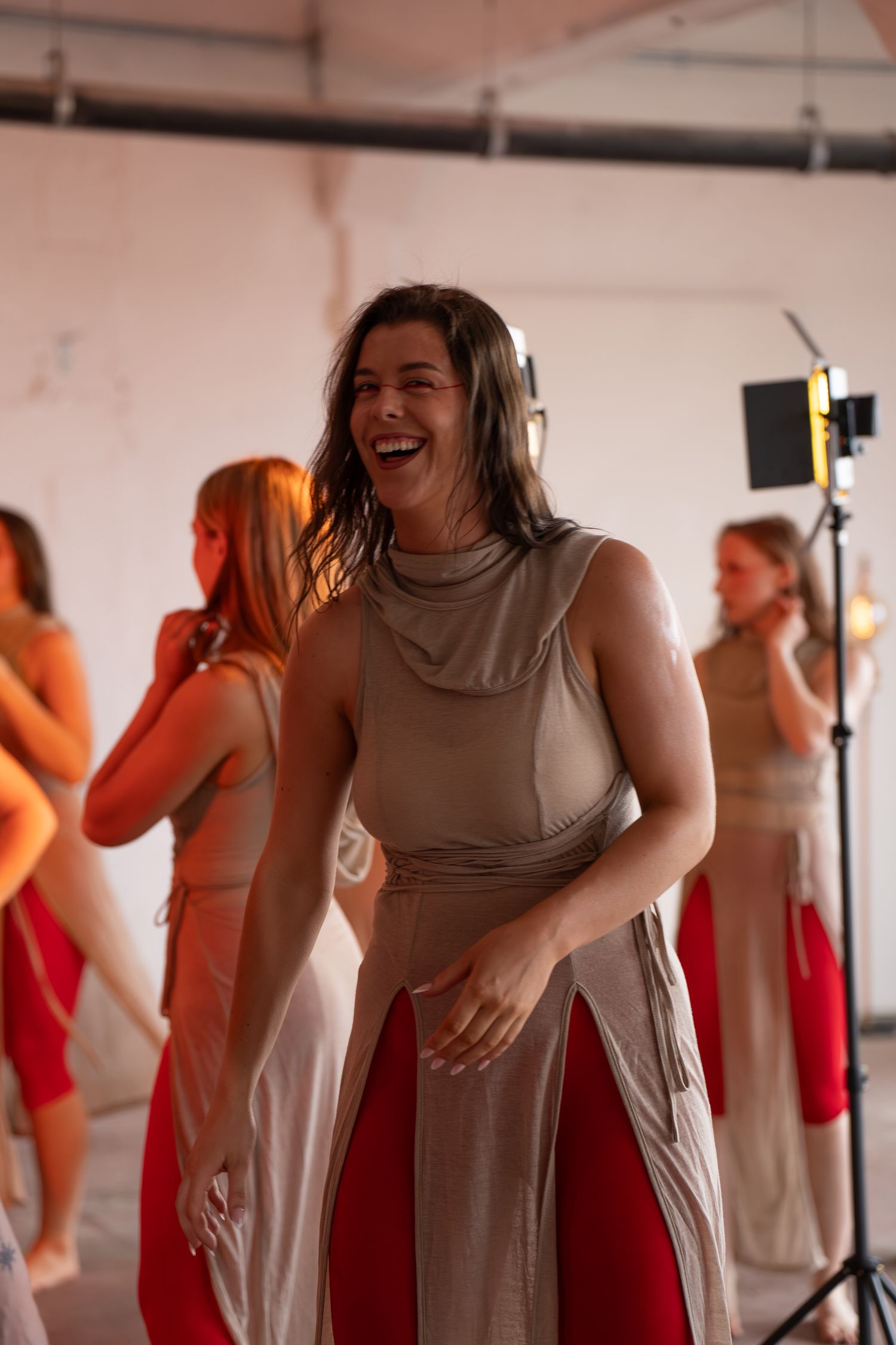 Smiling woman laughing in a beige tunic and red pants during a studio rehearsal with dancers and lighting equipment behind her.