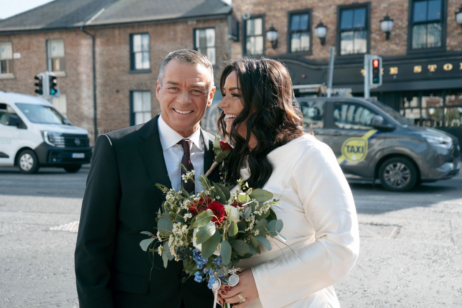 Smiling groom in a dark suit stands beside a bride in a white jacket holding a bouquet on a city street.