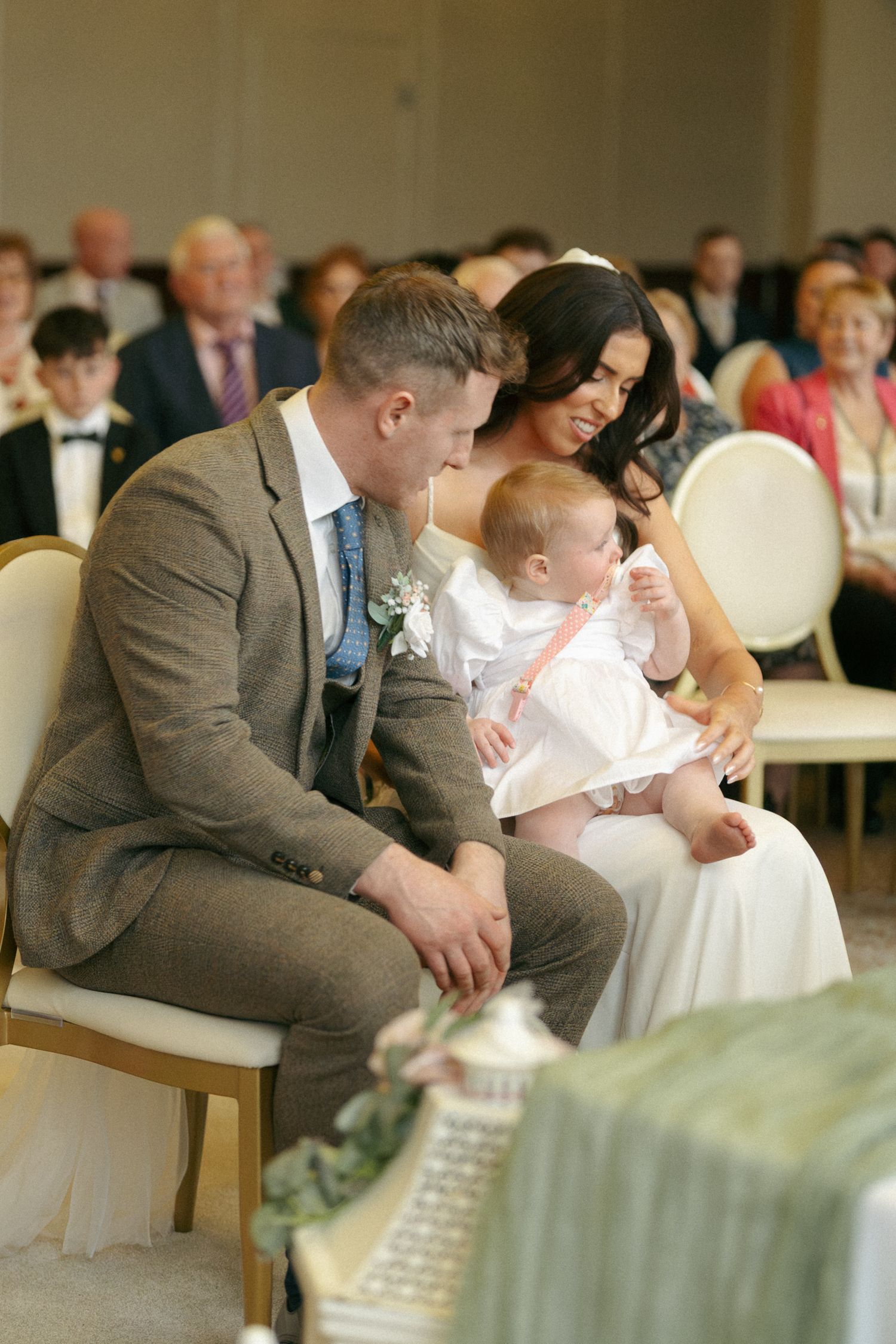 Groom in a tweed suit and bride in a white gown sit holding a baby during a wedding ceremony with guests behind them.