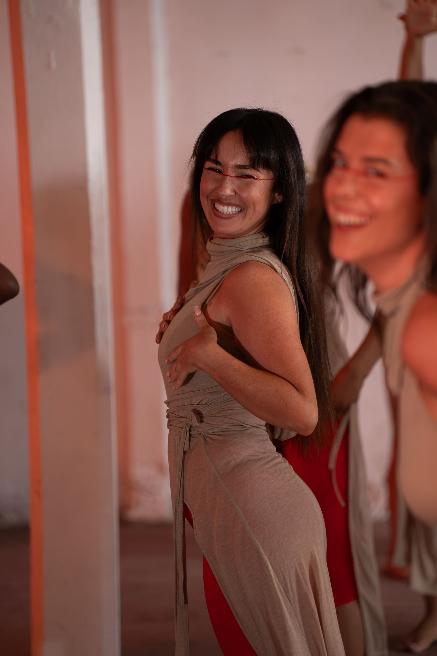 Smiling woman with long dark hair in a tan draped dress over red, dancing in a studio with another woman blurred behind her.