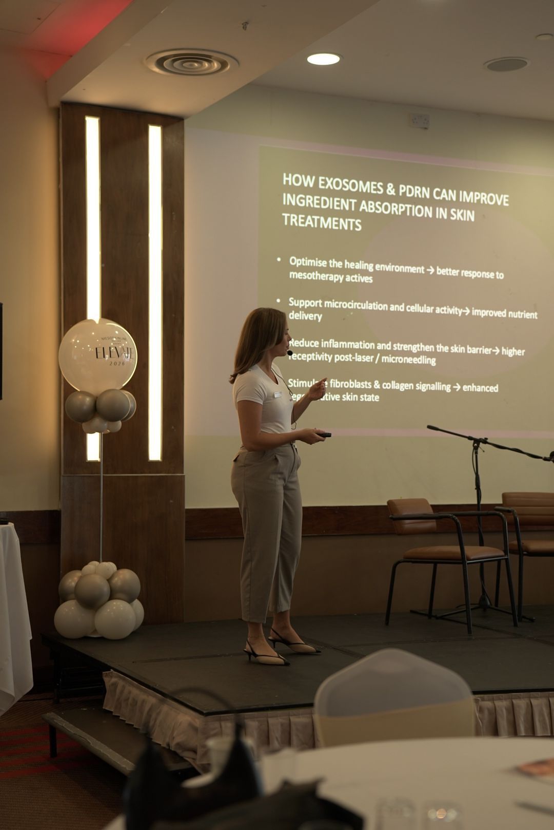 A presenter stands on stage beside a slide explaining how exosomes and PDRN can improve ingredient absorption in skin treatments.