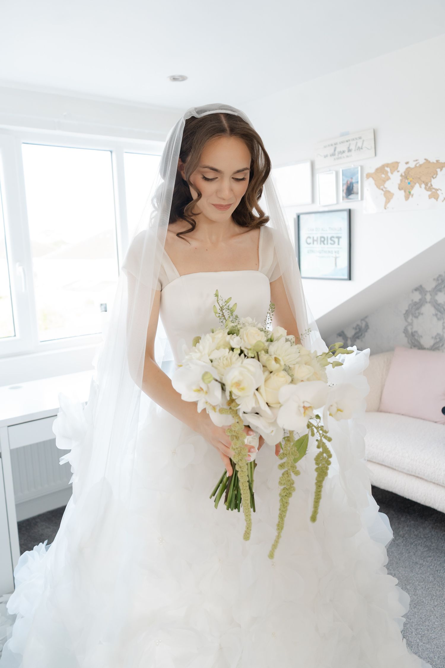 A bride in a ruffled white wedding gown and veil looks down at her white orchid bouquet with trailing green flowers in a bright room.
