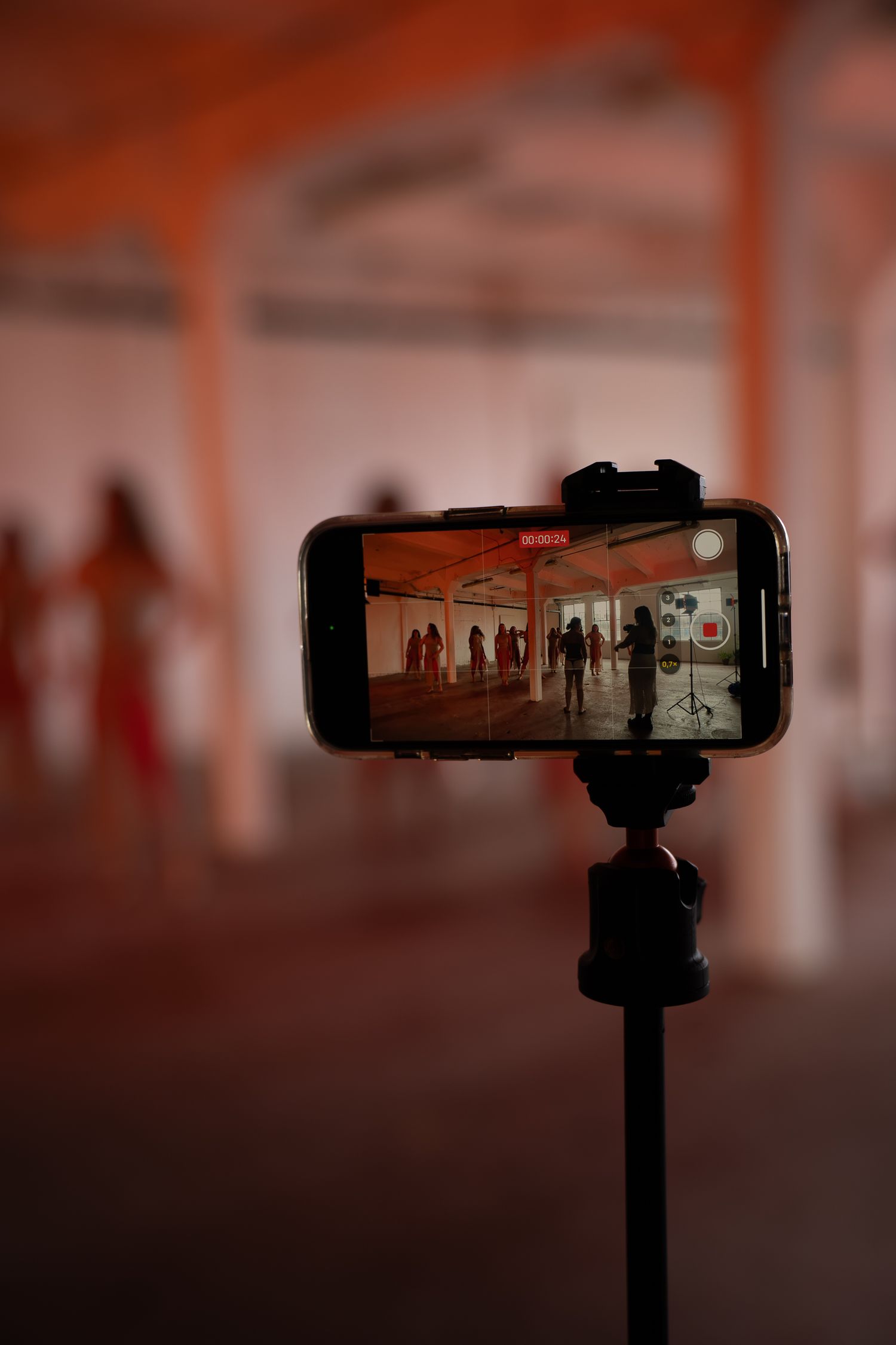 Smartphone on a tripod records people in red outfits standing in a dim, spacious hall with orange pillars.