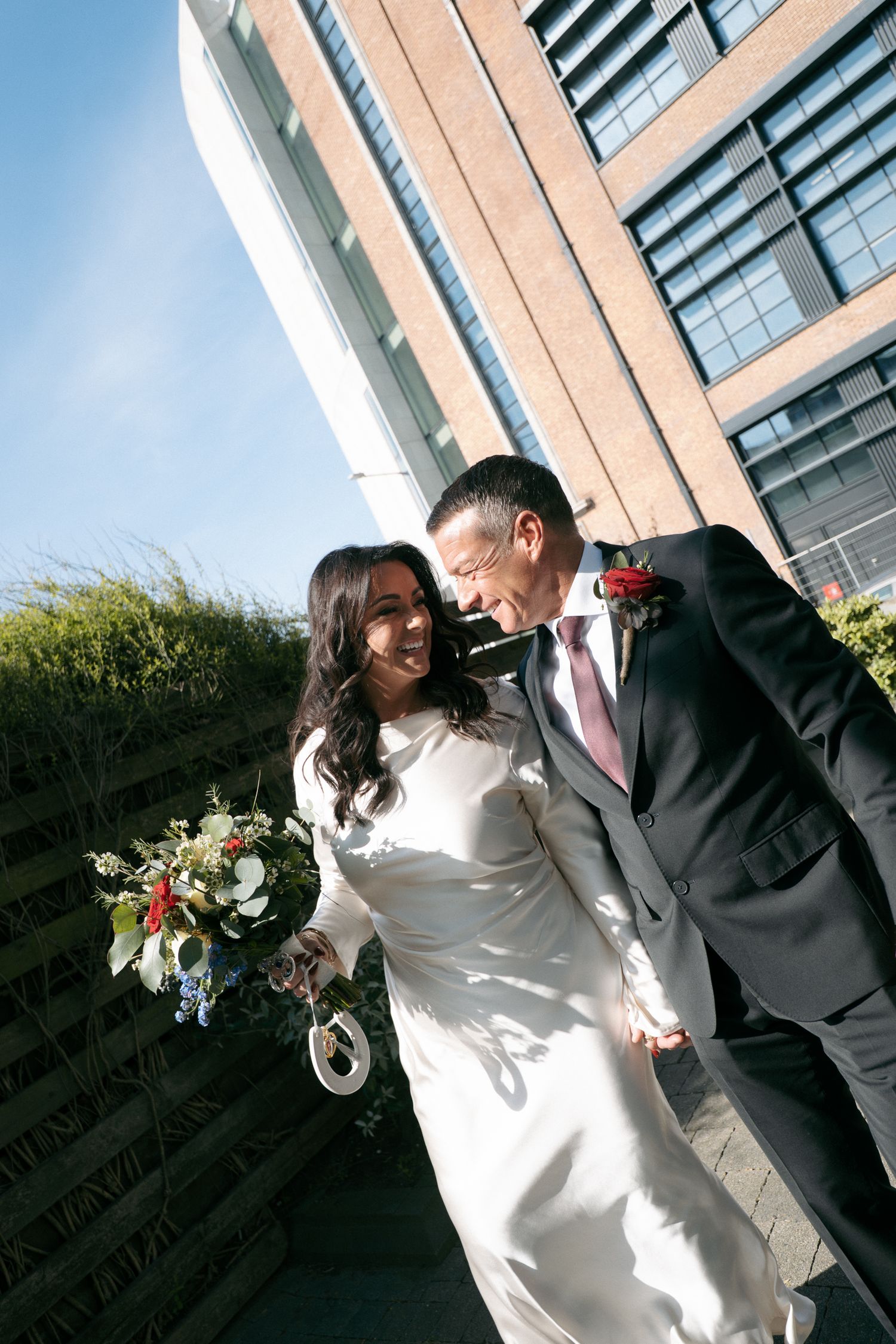 Smiling bride and groom walk past a modern brick and glass building, the bride holding a colorful bouquet and wearing a white wedding gown.