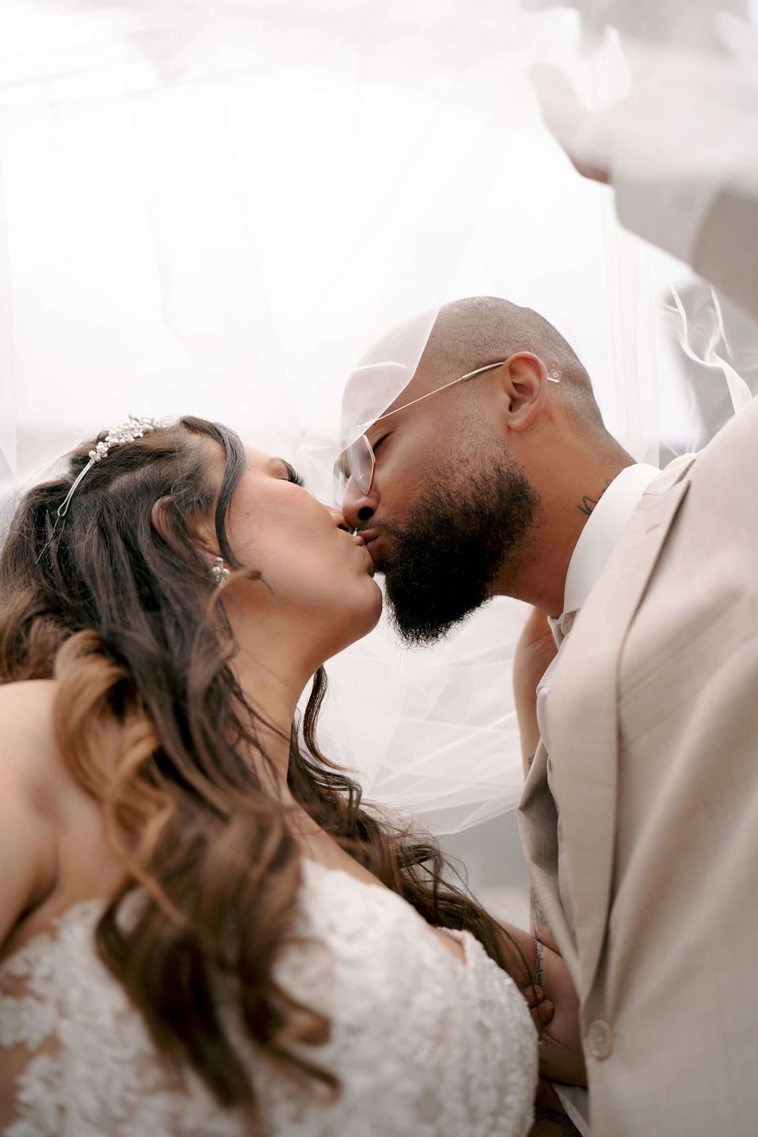 Bride in a lace wedding dress and groom in a beige suit and glasses share a kiss beneath a flowing sheer veil.