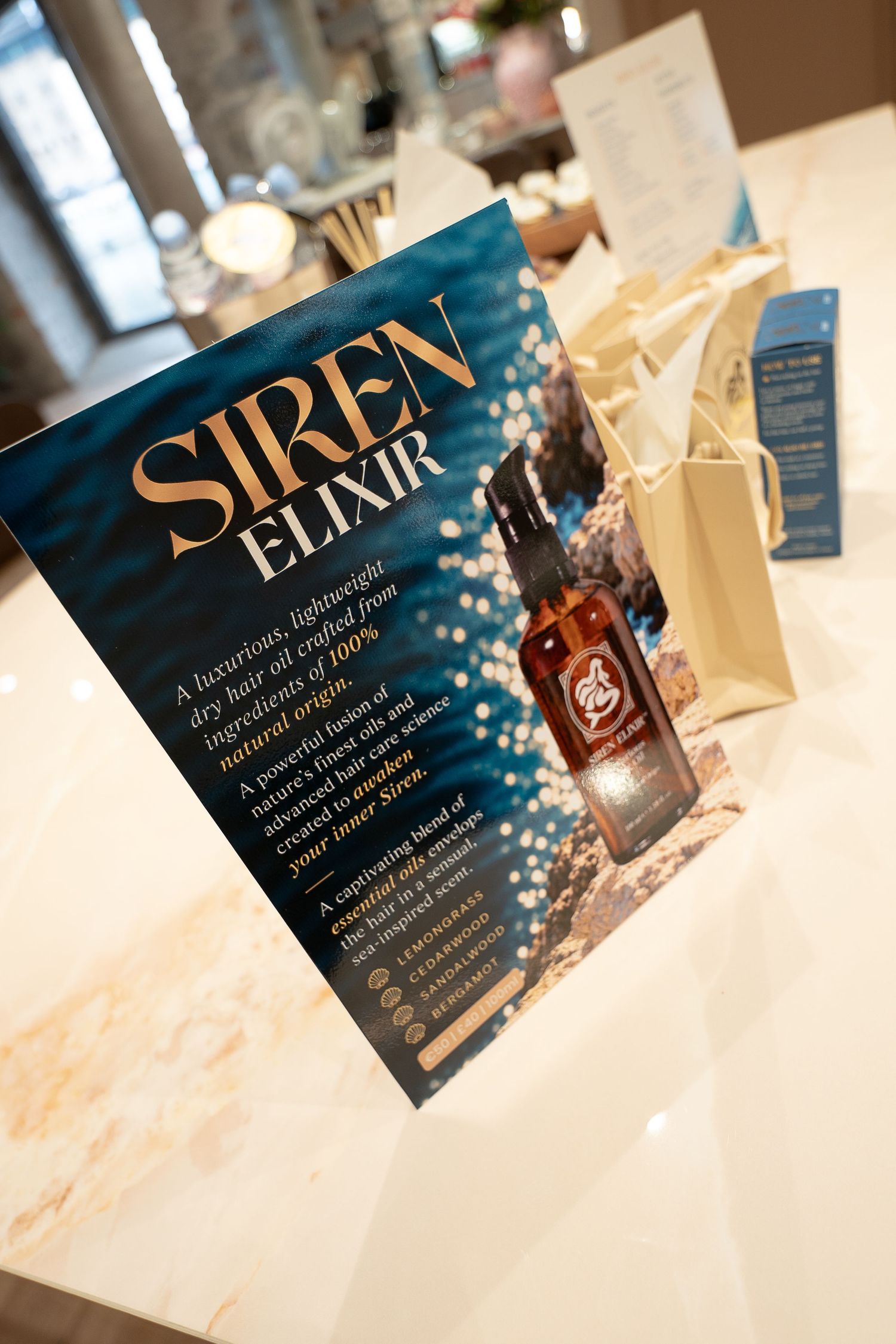Siren Elixir promotional display card with a dry hair oil bottle and cream gift bags on a marble salon counter.