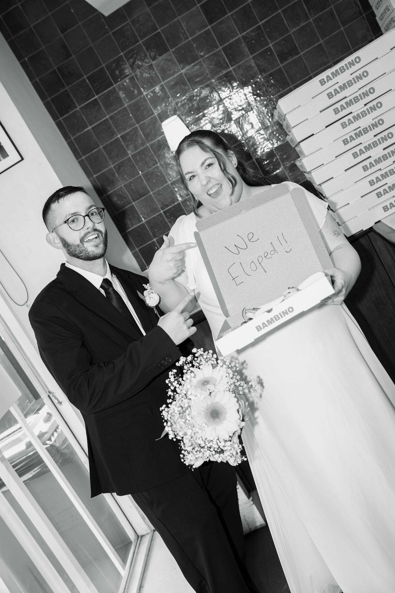 Smiling bride in a veil holds a Bambino pizza box reading We Eloped! beside her groom in glasses, black and white wedding photo.