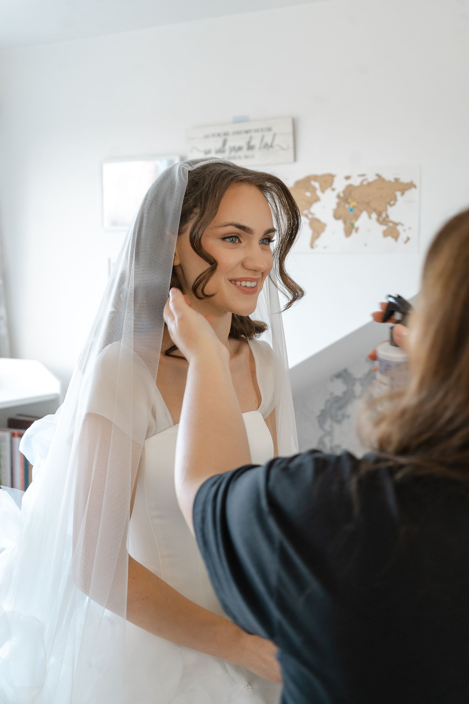 A smiling bride in a wedding gown and long veil has her hair styled by a stylist in a bright room with a world map on the wall.