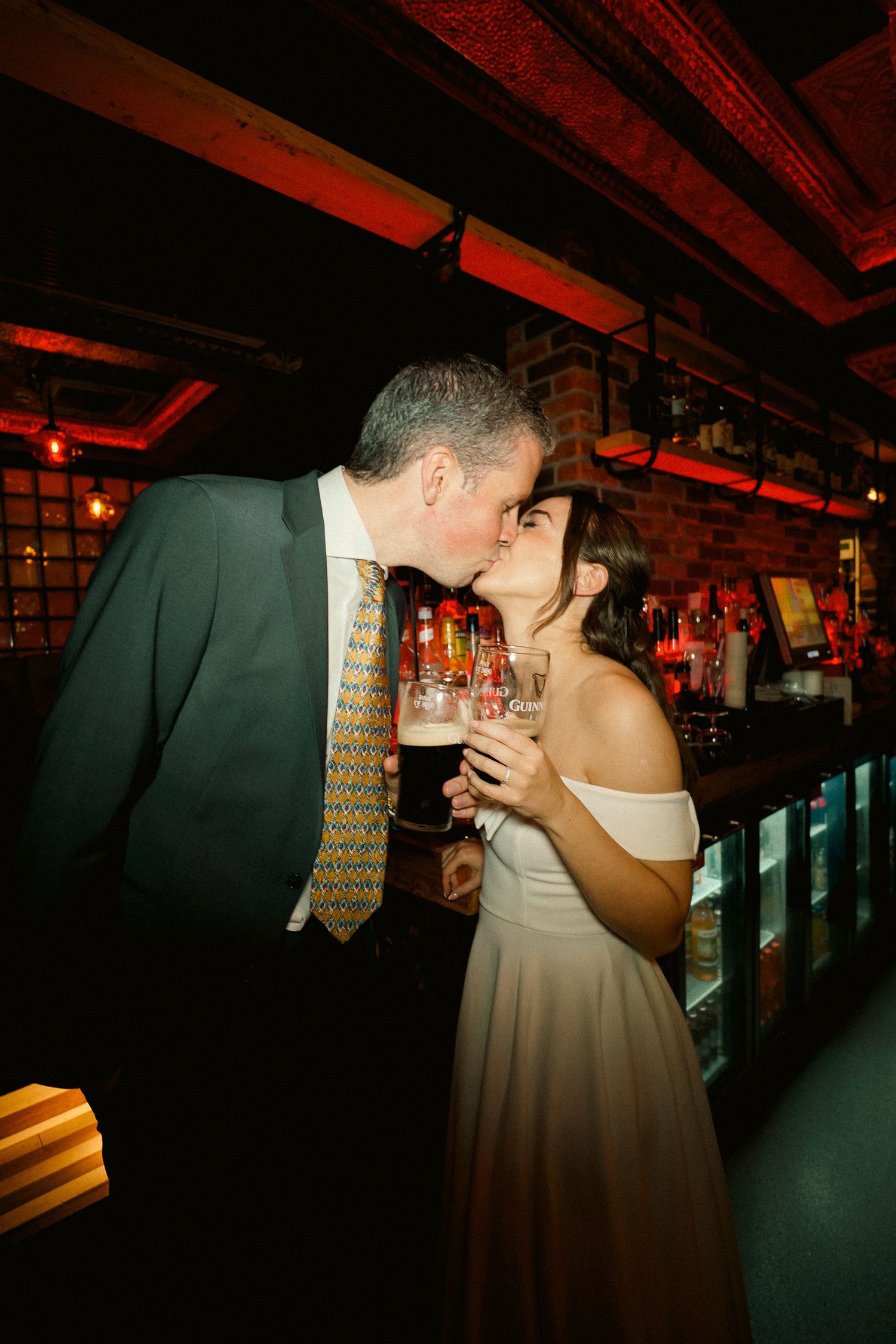 Bride and groom kiss at a moody pub bar while holding pints of stout after their wedding.