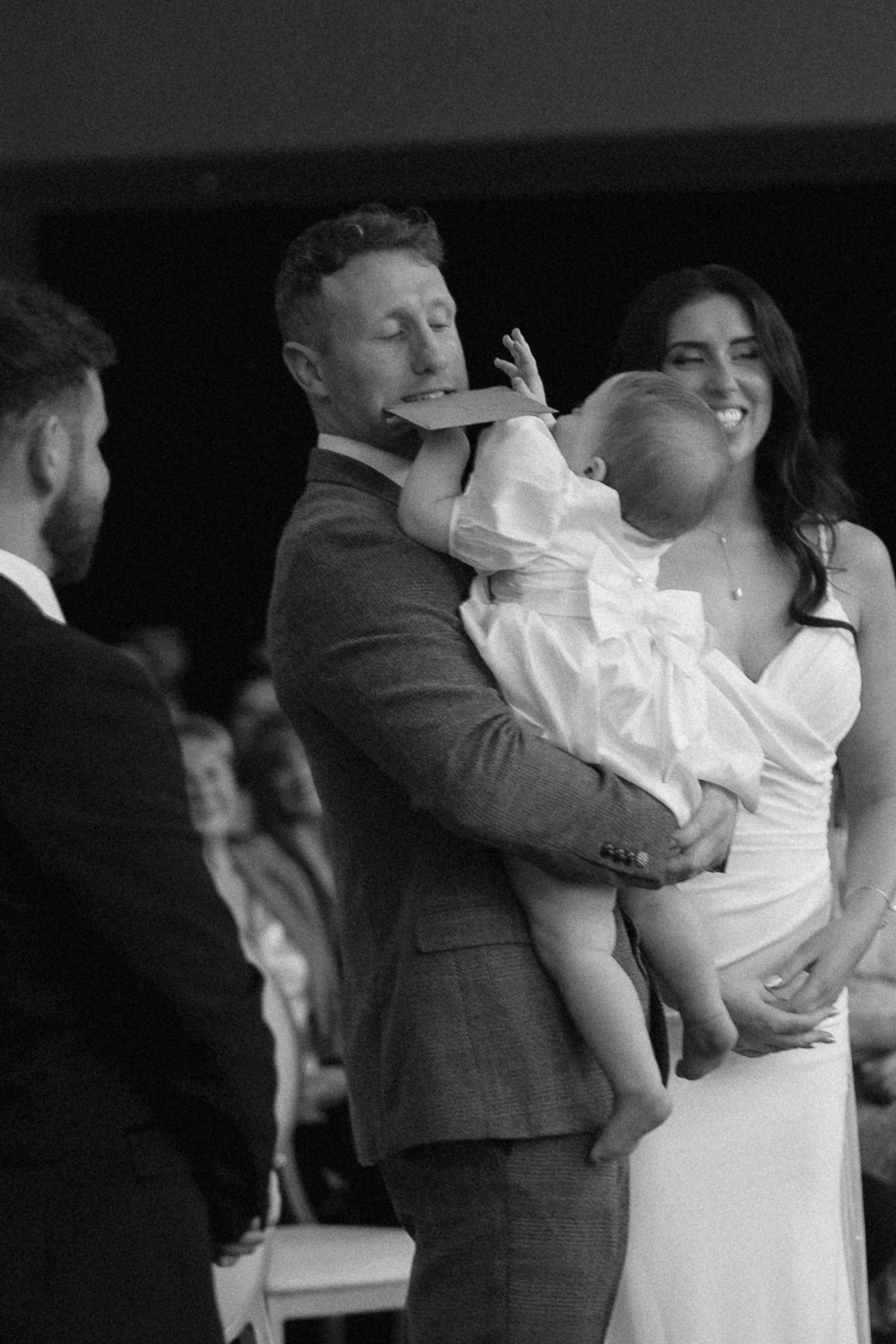 Black and white photo of a smiling bride beside a groom holding a baby in a white dress during a wedding ceremony.