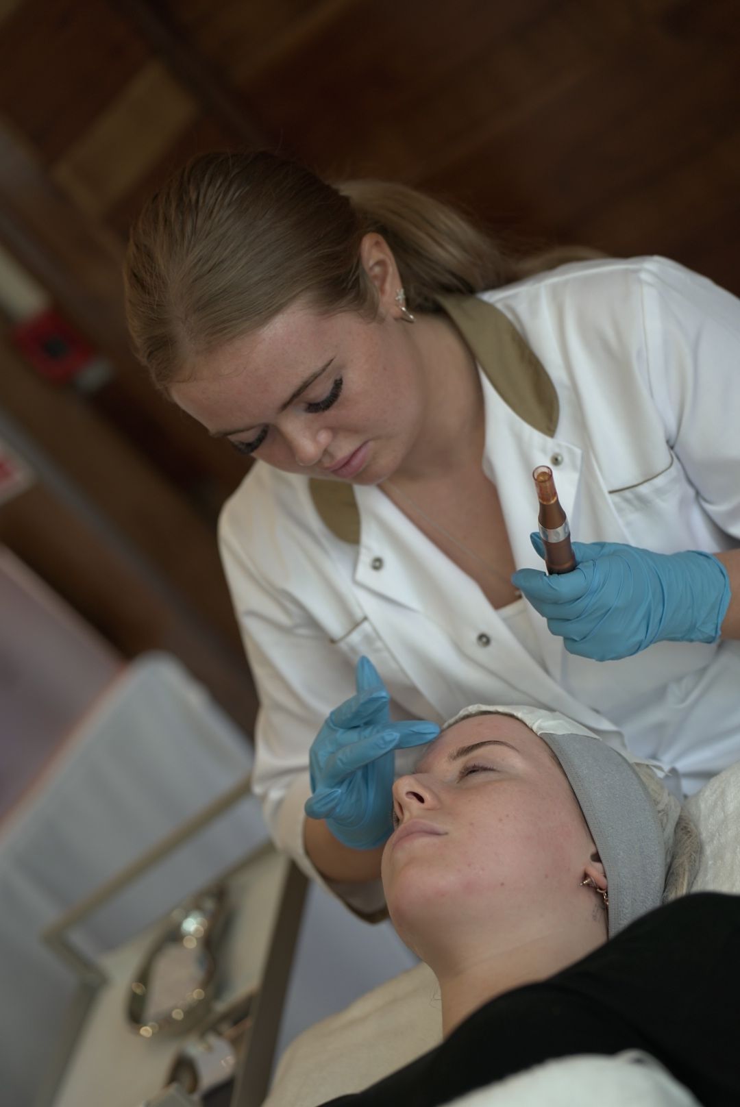 A woman in a white tunic and blue gloves applies a serum to a client's forehead during a facial treatment at a beauty event.