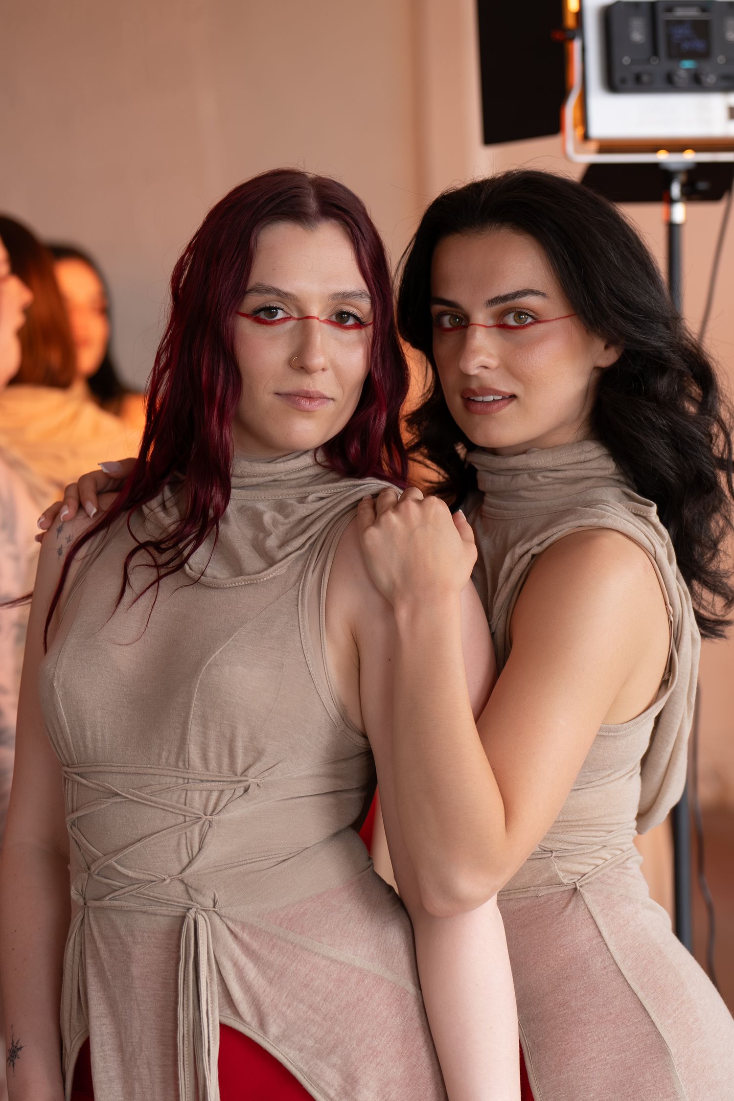 Two women with red and dark hair pose closely in matching beige cowl-neck lace-up tops during a studio photo shoot with lighting equipment.