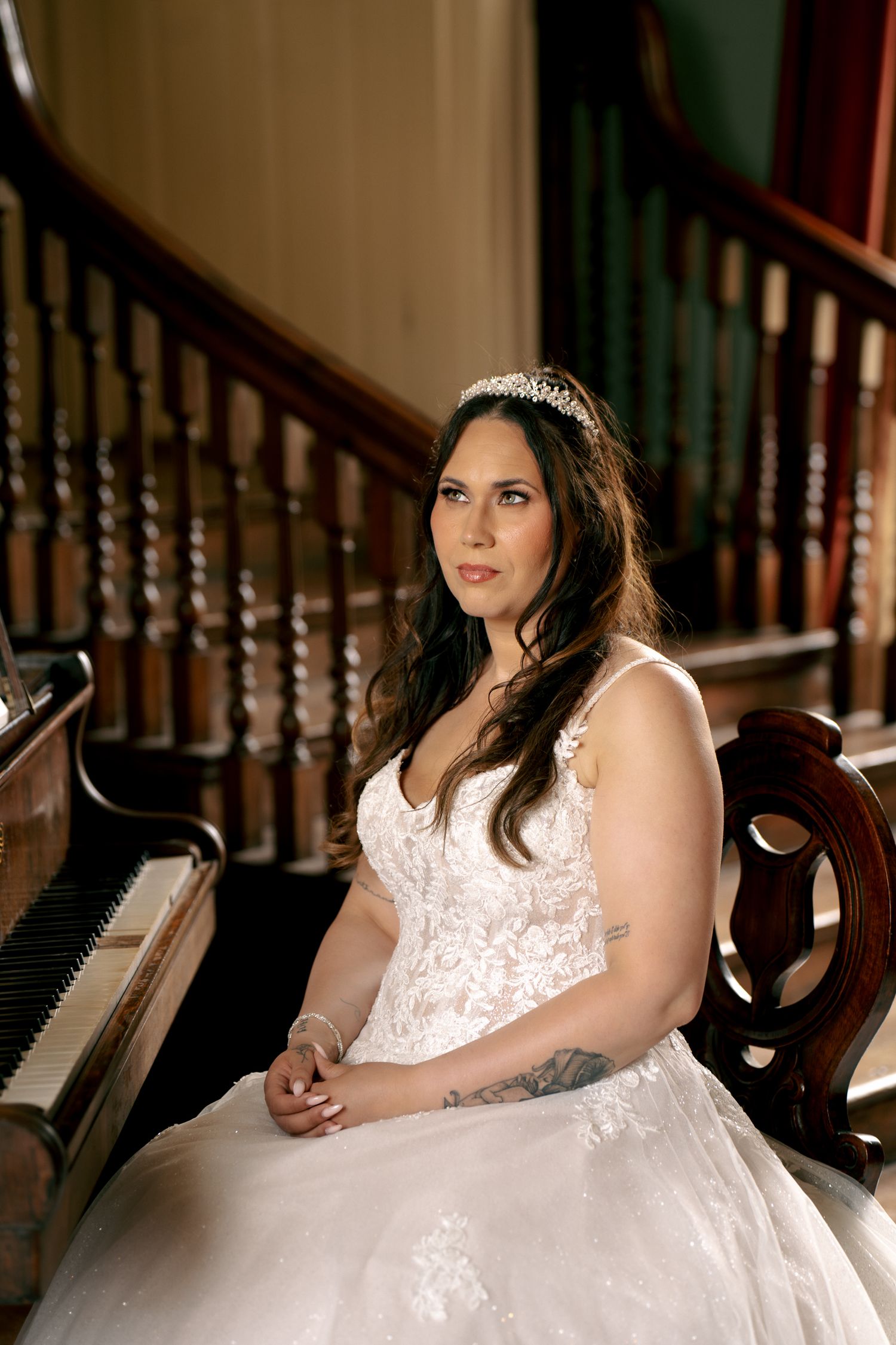 A bride wearing a lace gown and sparkling tiara sits thoughtfully near a piano and wooden staircase in a historic interior.