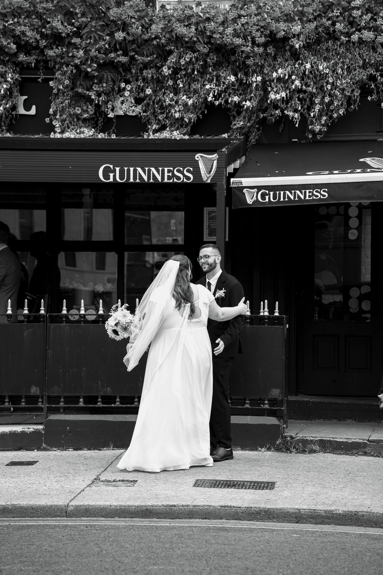 Bride in a long veil and groom in a dark suit embrace on the street outside a Guinness pub with ivy overhead, black and white photo.