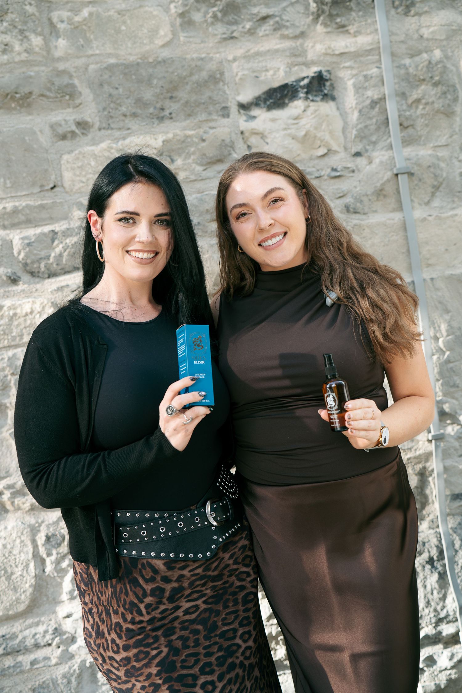 Two smiling women stand against a stone wall holding Siren Elixir hair oil products, one with a blue box and one with a bottle.
