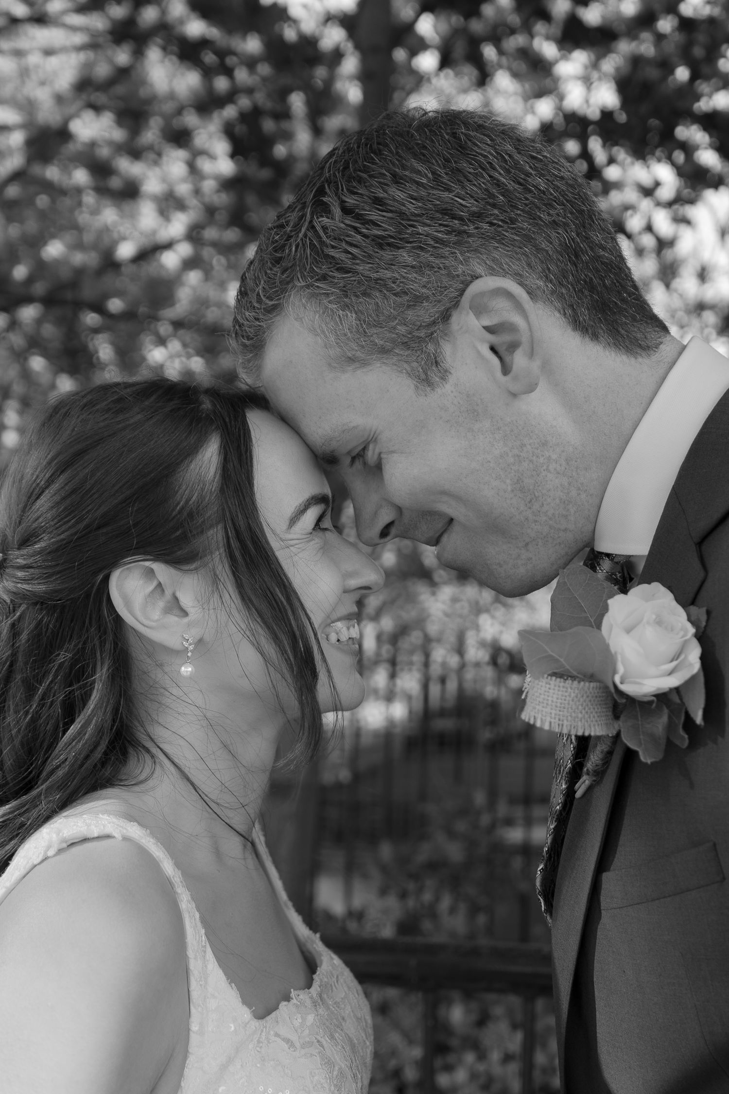 Black and white portrait of a smiling bride and groom touching foreheads, the groom wearing a rose boutonniere, outdoors among trees.