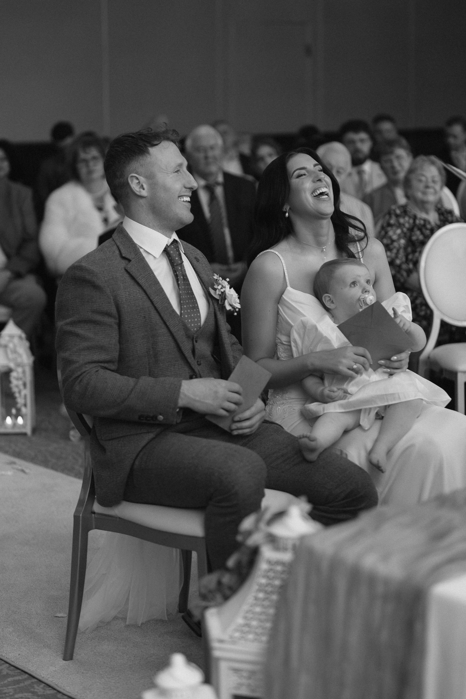 Black and white photo of a laughing couple holding a baby during a wedding ceremony with guests seated behind.