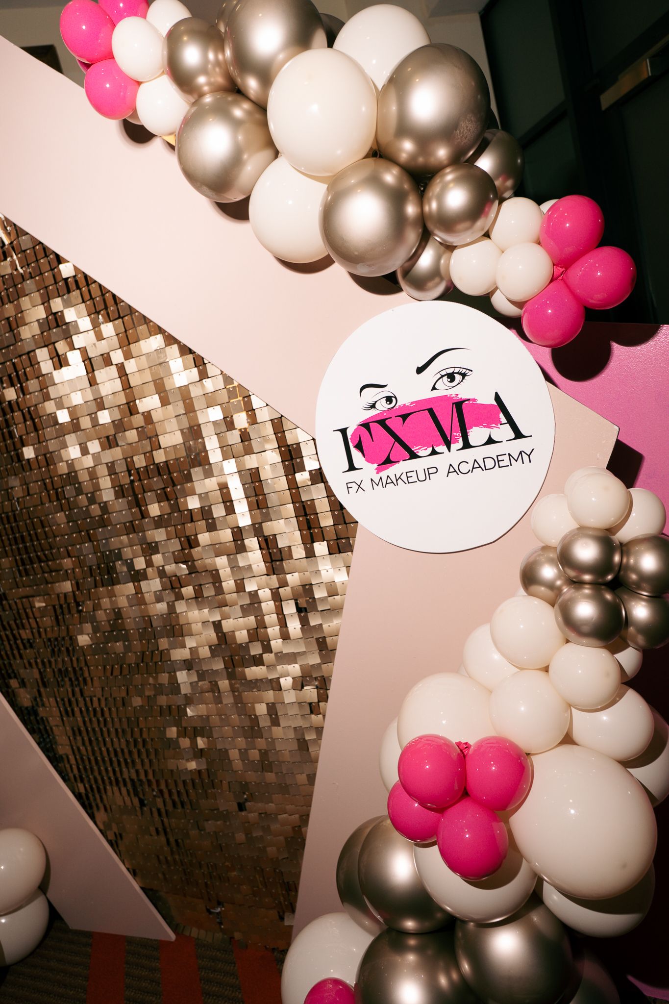 A pink, white and champagne balloon garland frames an FX Makeup Academy logo beside a sparkling gold sequin wall.