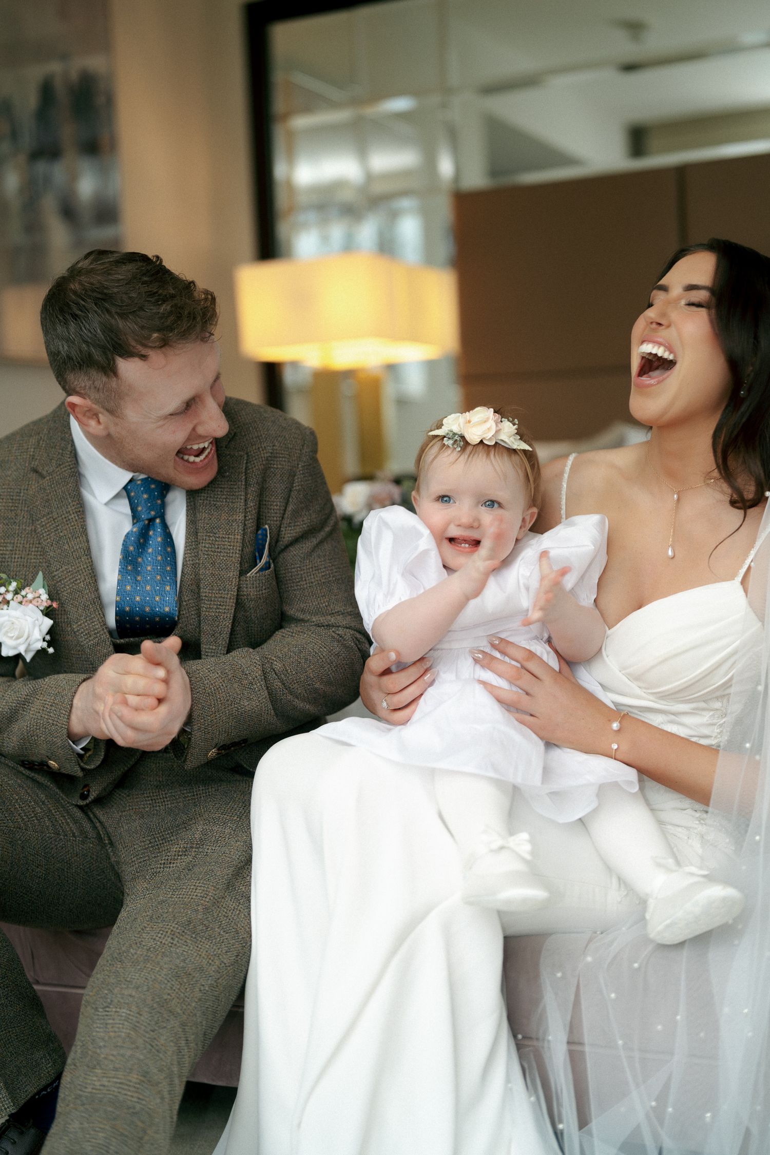 Laughing bride and groom sit together on a sofa with a baby girl in a white dress and floral headband.