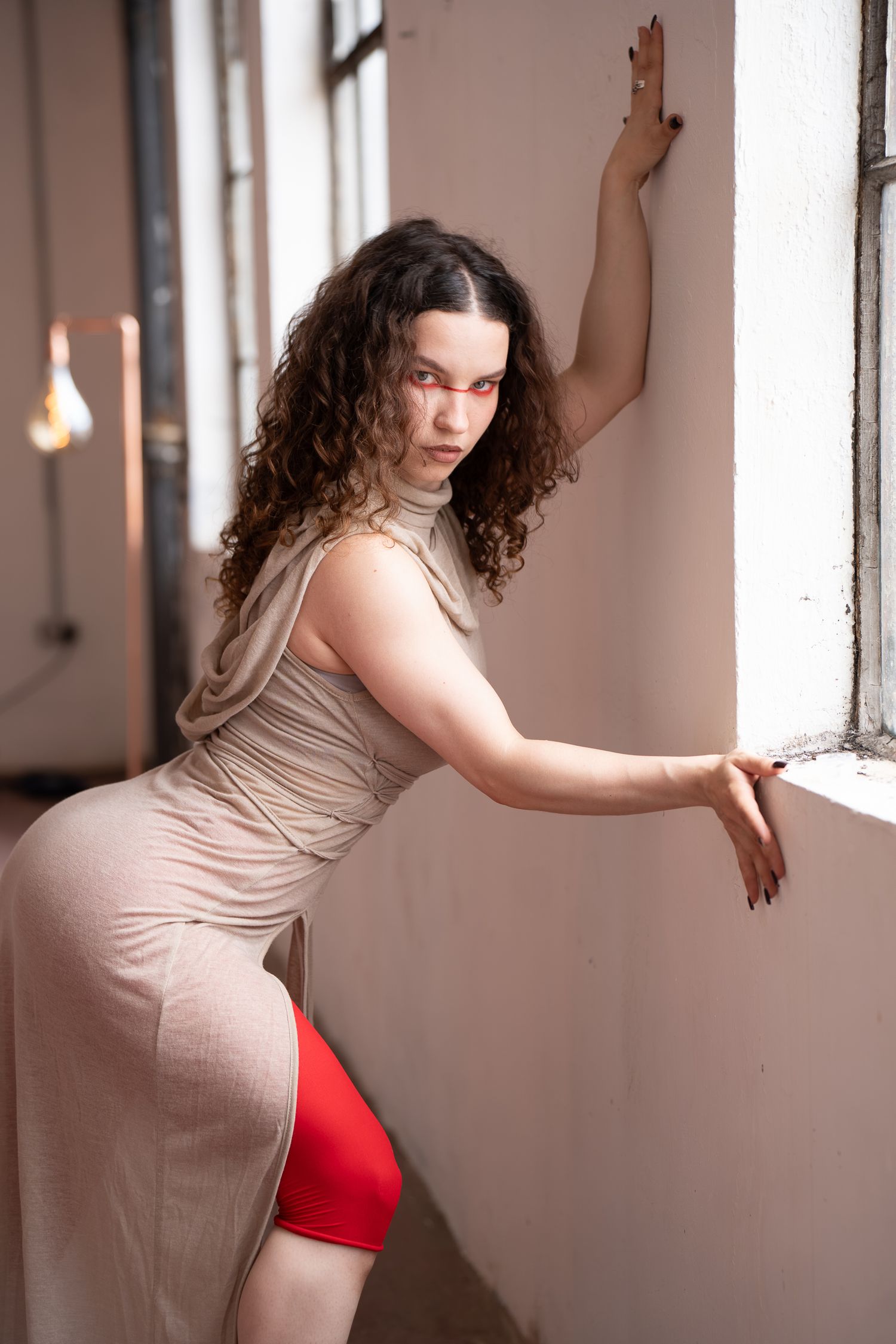 Woman with long curly dark hair and red eye makeup poses in a beige draped dress and red shorts, leaning against a sunlit studio wall.