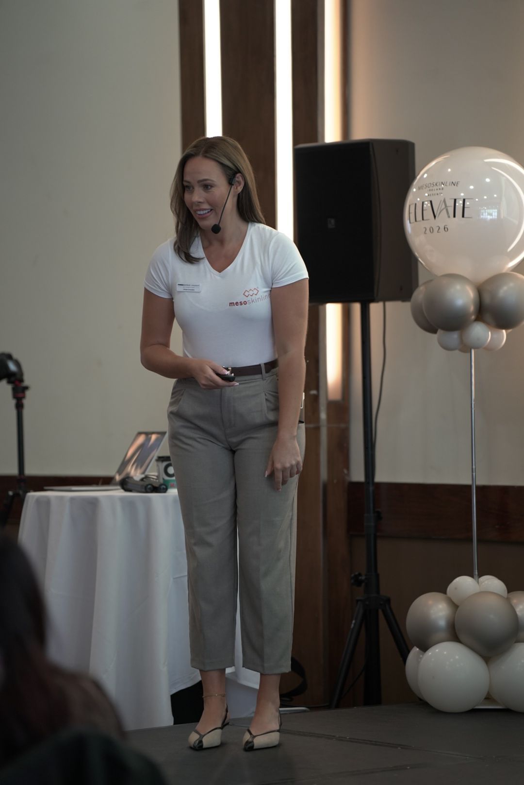 A woman with a headset microphone stands on stage near a speaker and balloon display at the Elevate skincare conference.