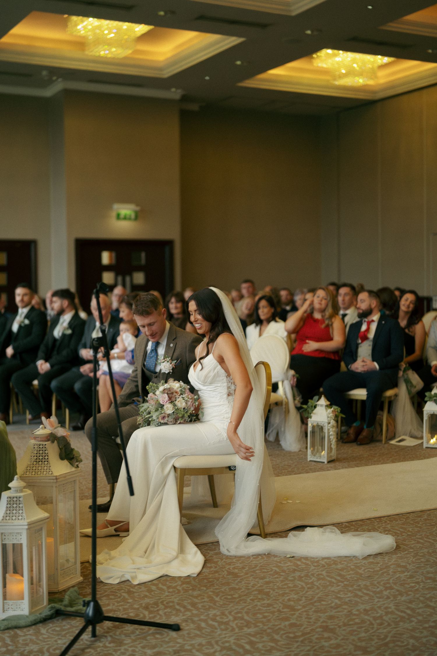Bride in a white gown with a long veil sits holding a bouquet in a ballroom ceremony with guests seated in rows.