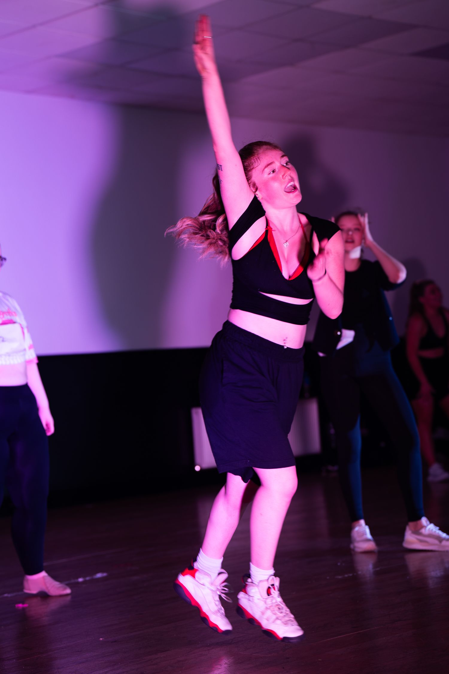 A dancer in a black crop top and skirt leaps mid-motion with one arm raised during a dance performance under pink stage lighting.
