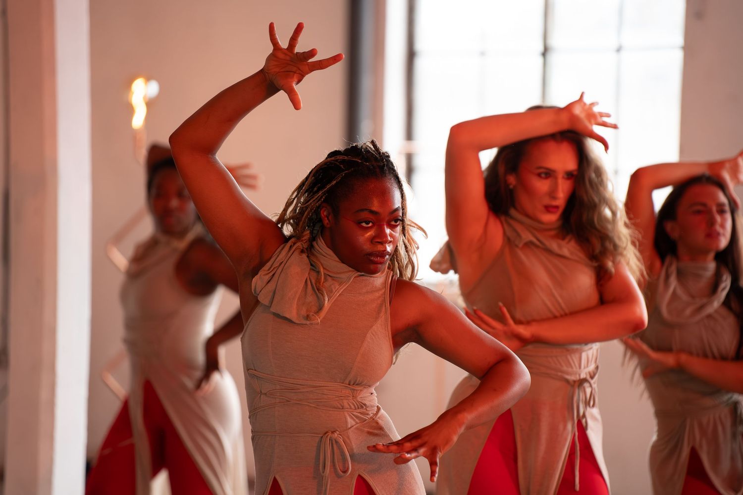 Group of dancers in matching beige tops and red pants strike expressive poses with raised arms in a bright, sunlit studio.