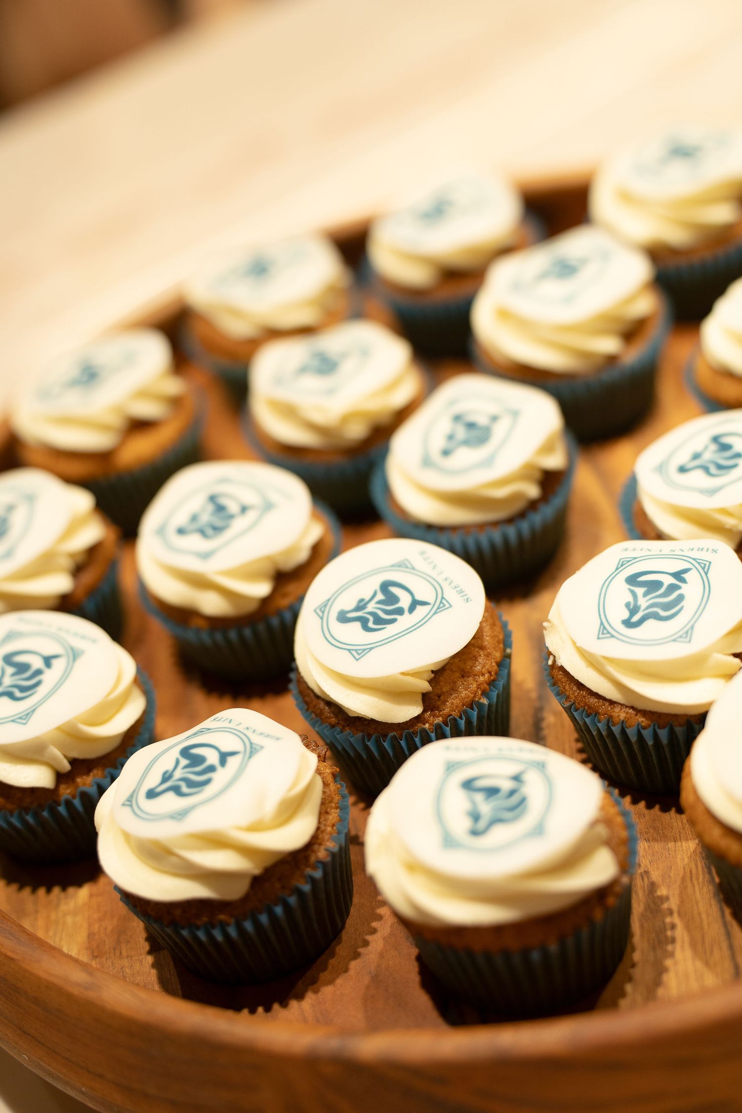 Tray of chocolate cupcakes topped with white buttercream and edible logo discs arranged on a round wooden serving board.