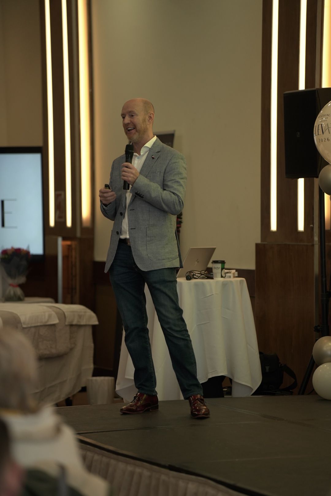 A bald man in a gray blazer and jeans speaks into a microphone on stage at a beauty industry conference.