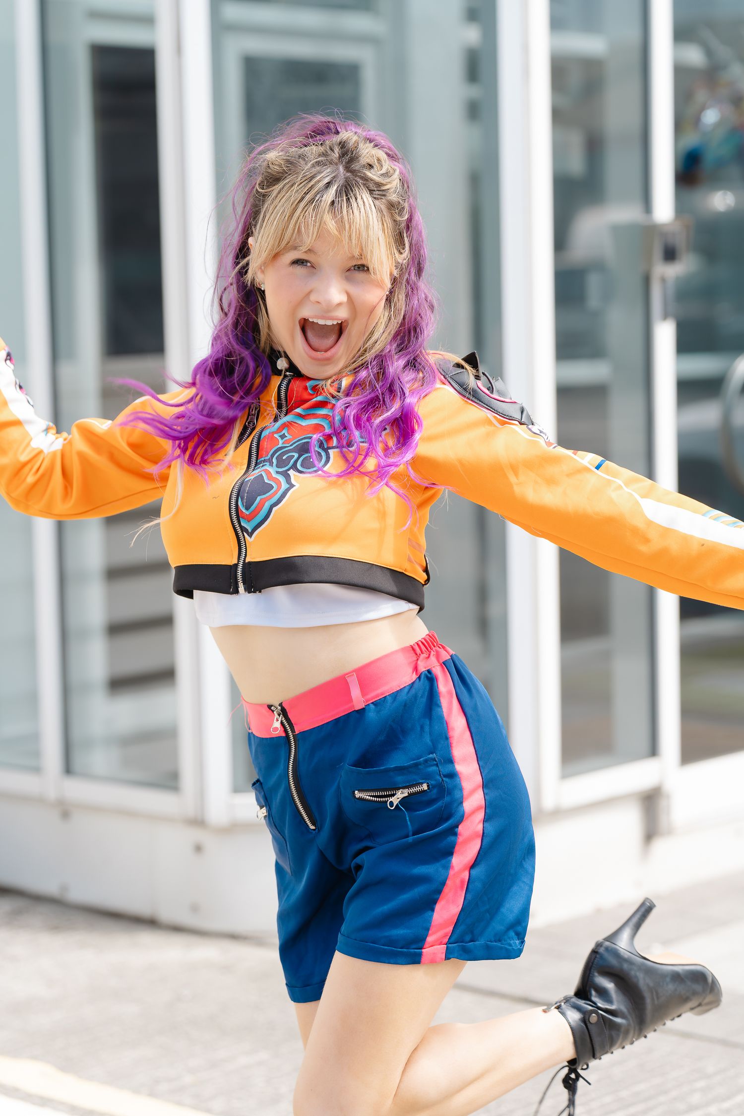 Woman with purple-dyed hair laughs with arms outstretched in an orange racing jacket, blue shorts and black boots outside a glass building.