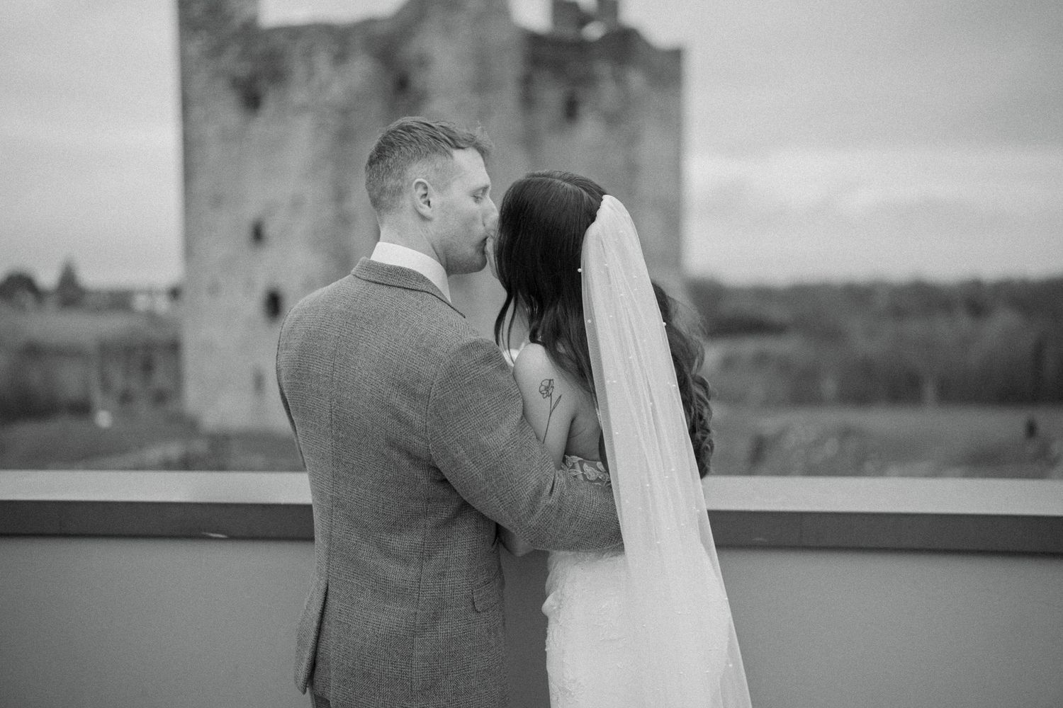 Black and white photo of a groom kissing his bride in a long veil on a terrace overlooking a castle.