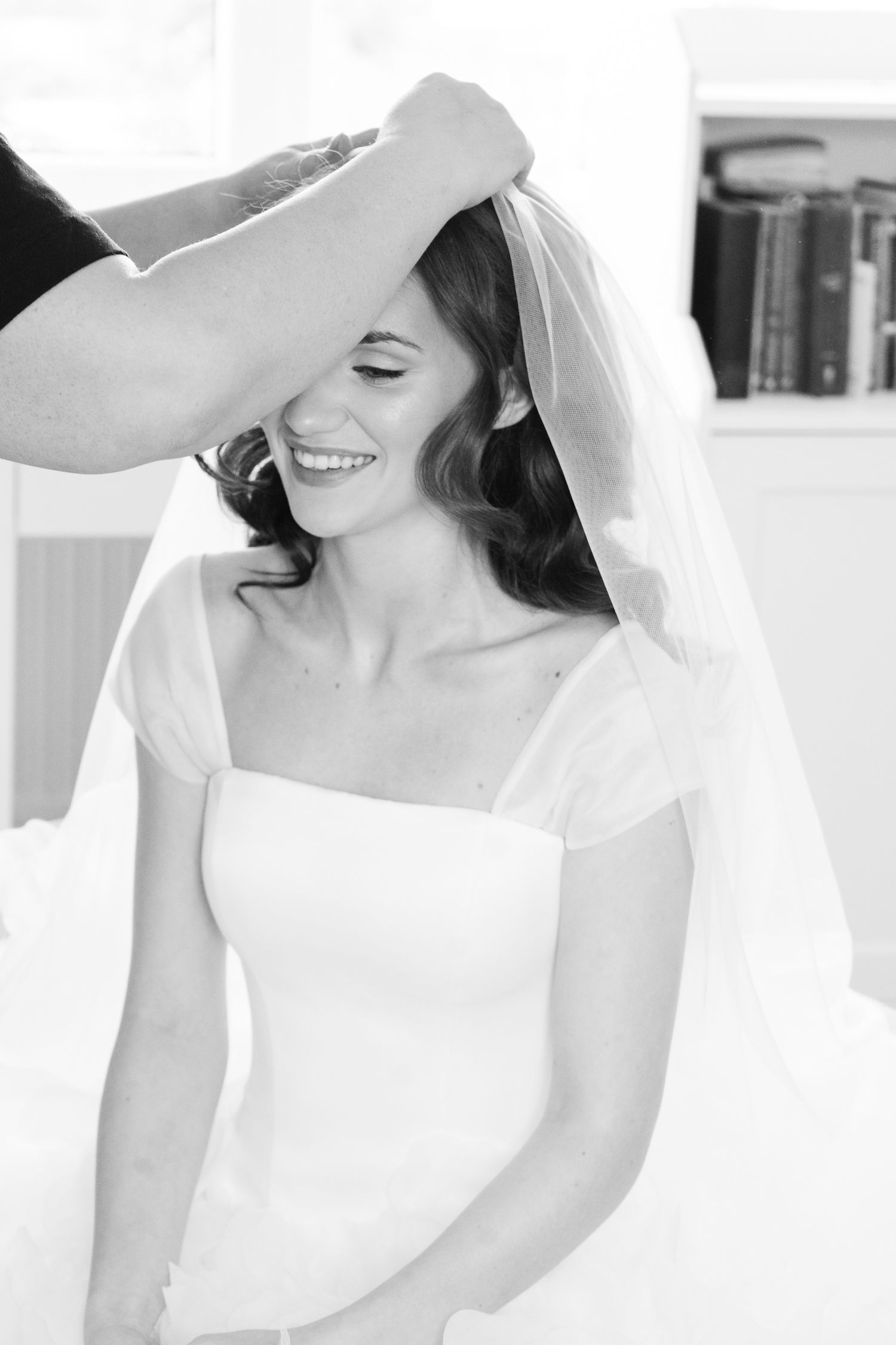 Black and white photo of a smiling bride in an off-shoulder gown as a stylist adjusts her sheer wedding veil.