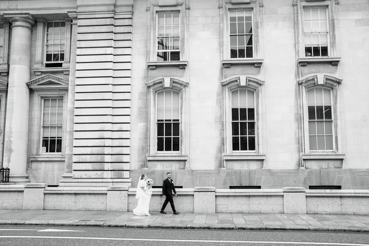 Bride in a long white gown and groom in a dark suit walk past a grand stone building with tall windows in a black and white photo.