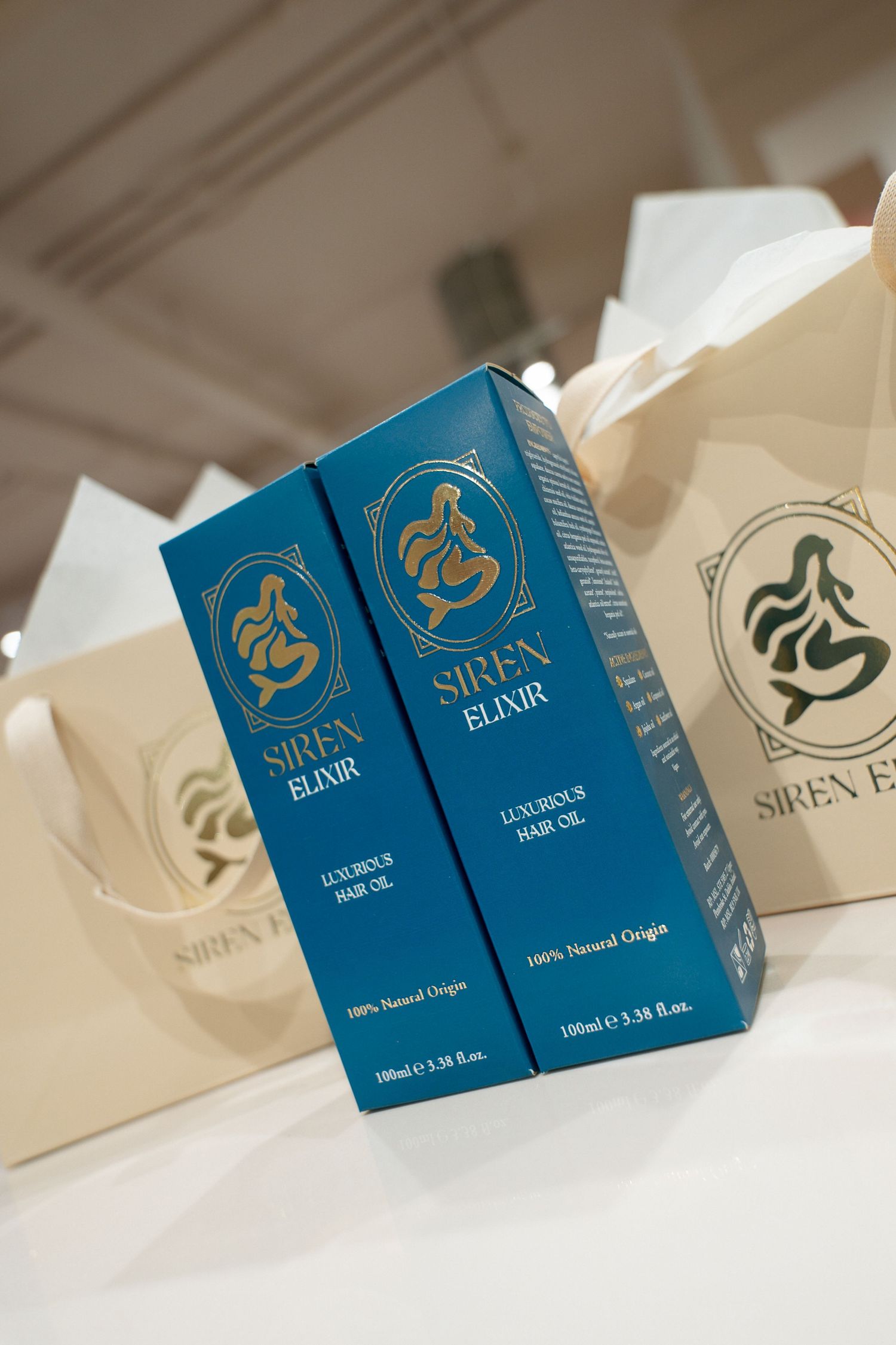 Two blue Siren Elixir luxurious hair oil boxes with gold mermaid logos beside cream branded gift bags.