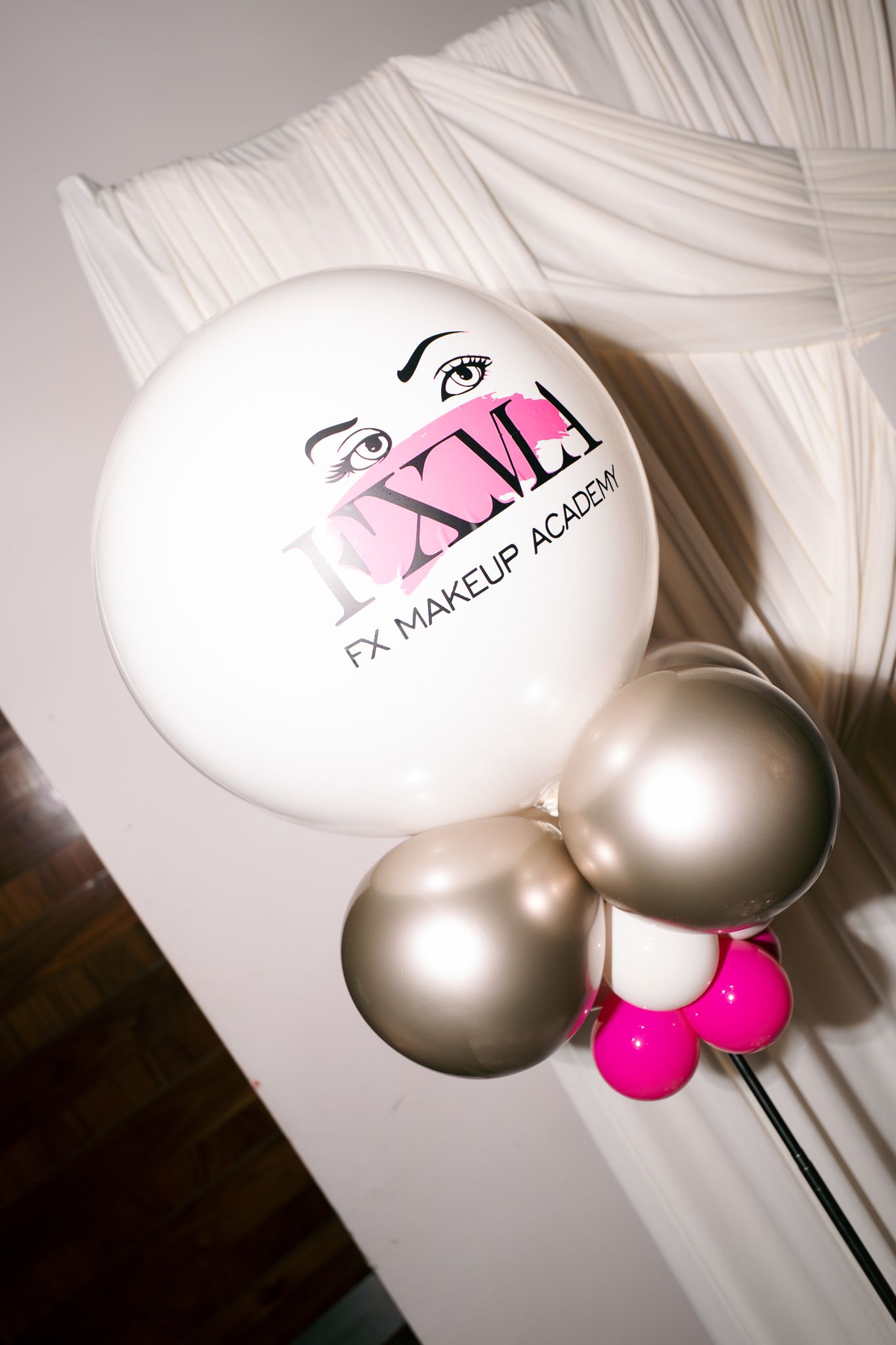 White FX Makeup Academy balloon with eyes logo, tied to silver and hot pink balloons, displayed beside white draped fabric at an event.
