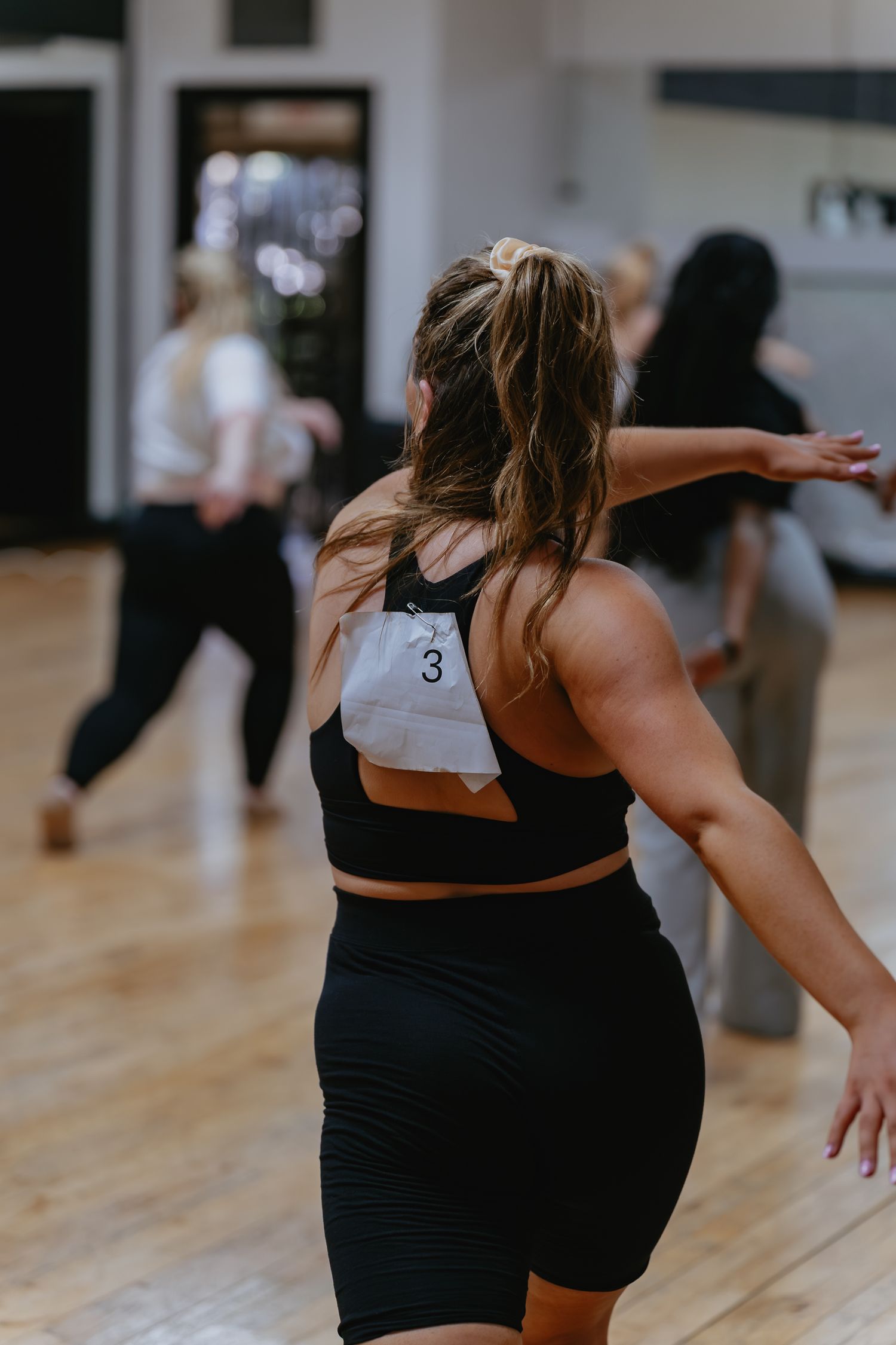Dancer with a ponytail and number 3 pinned to her black athletic outfit performs in a studio with other dancers on a wood floor.