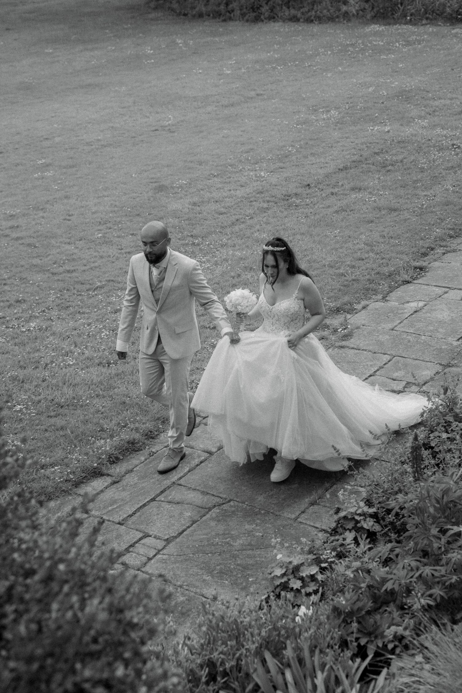 In black and white, a bride and groom walk hand in hand along a stone garden path beside a lawn, the bride holding a bouquet.