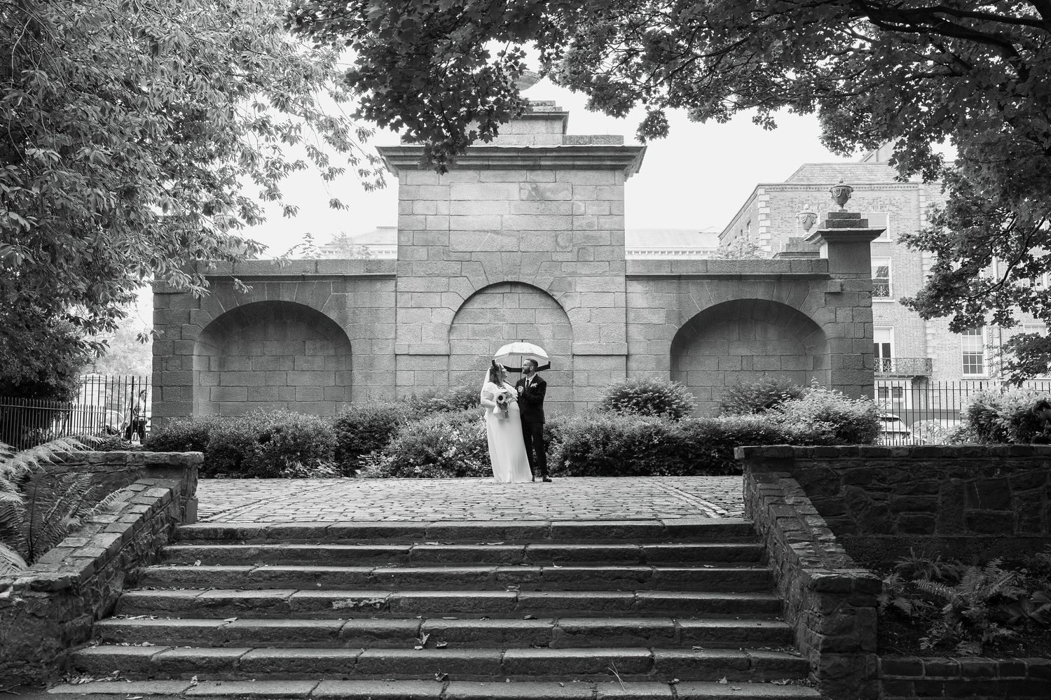 Newlyweds share an umbrella at the top of stone steps in front of an arched stone wall on a rainy day, black and white wedding photo.