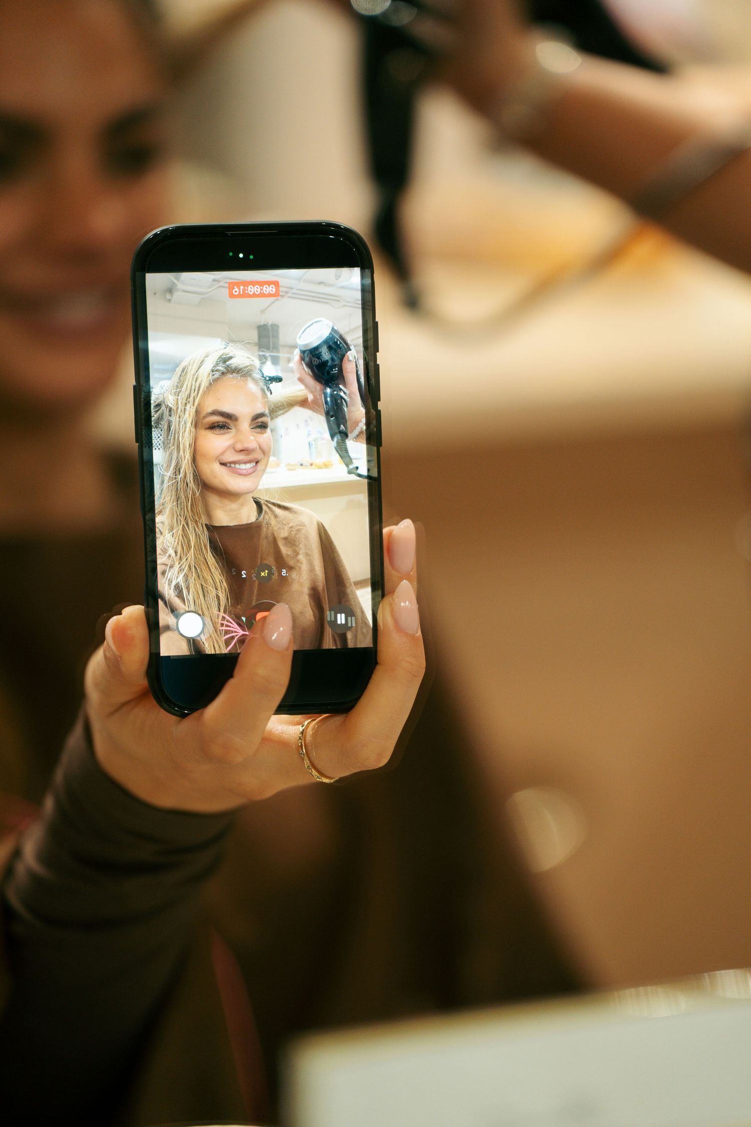 Hand holding a smartphone showing a smiling blonde woman filming a selfie video during a salon hair styling session.