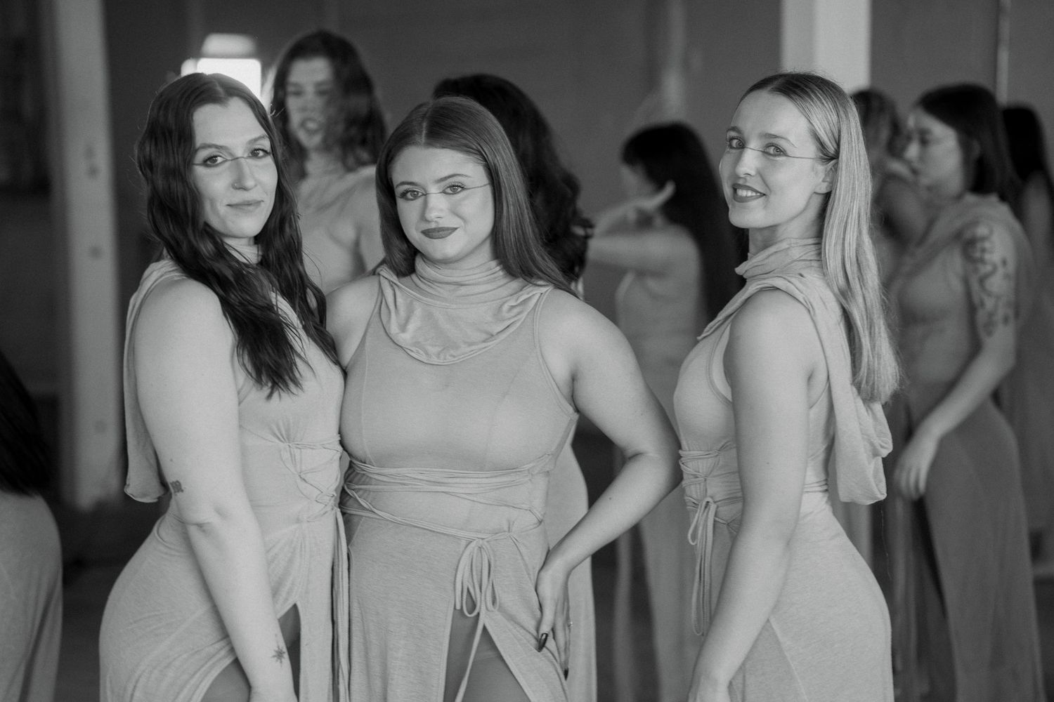 Black and white photo of three smiling bridesmaids in matching satin dresses posing close together with other bridesmaids behind them.