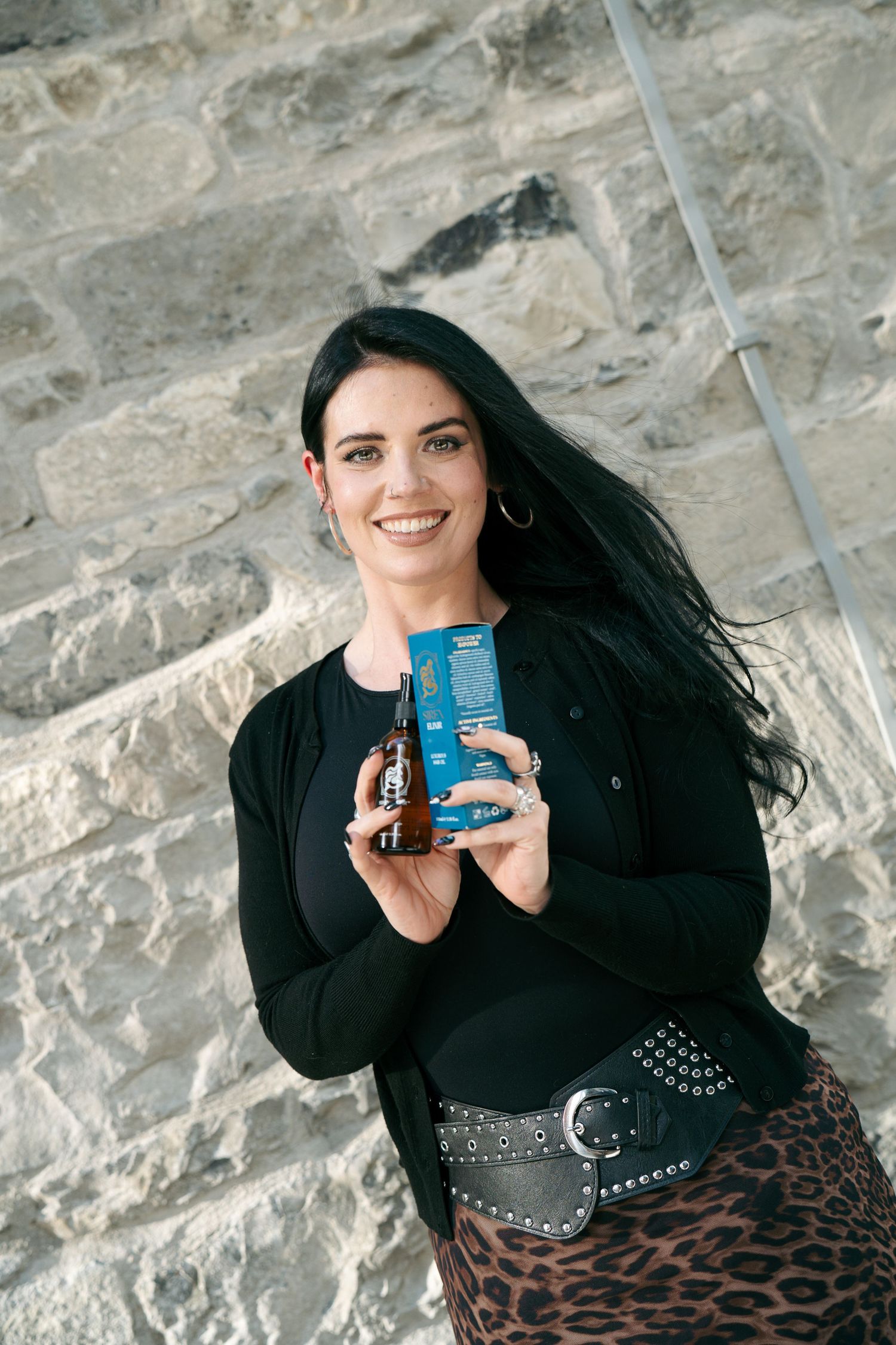 Smiling woman with long black hair in a black cardigan and leopard skirt holds a blue product box and amber bottle by a stone wall.