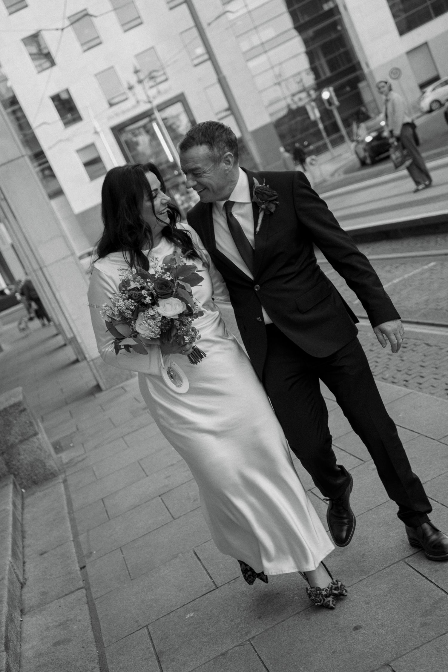 Black and white image of a laughing wedding couple strolling a city sidewalk, the bride holding a bouquet and leaning toward her groom.